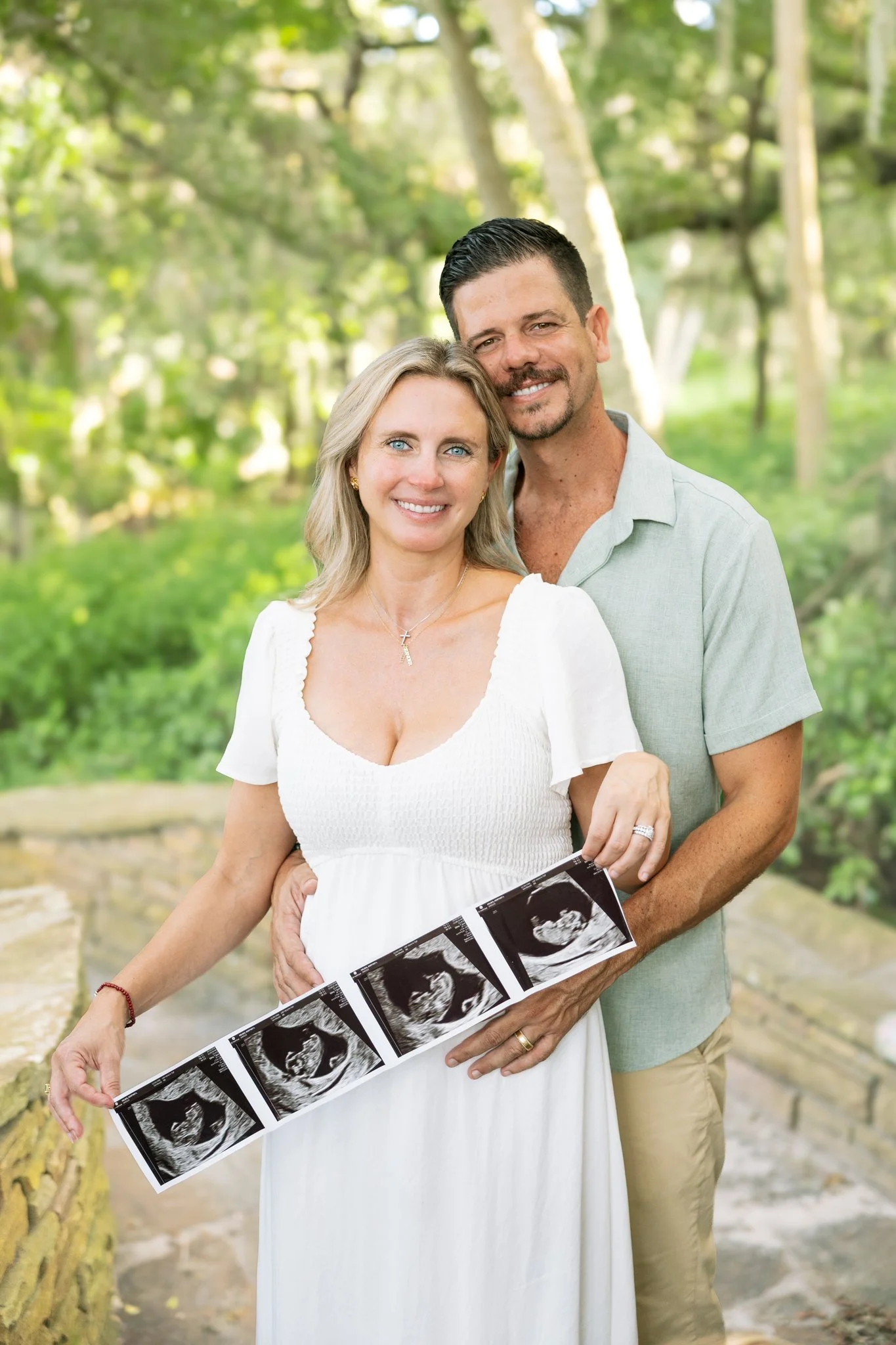 Couple holding an ultrasound strip on a shaded stone path at Philippe Park, his hand resting on the baby bump