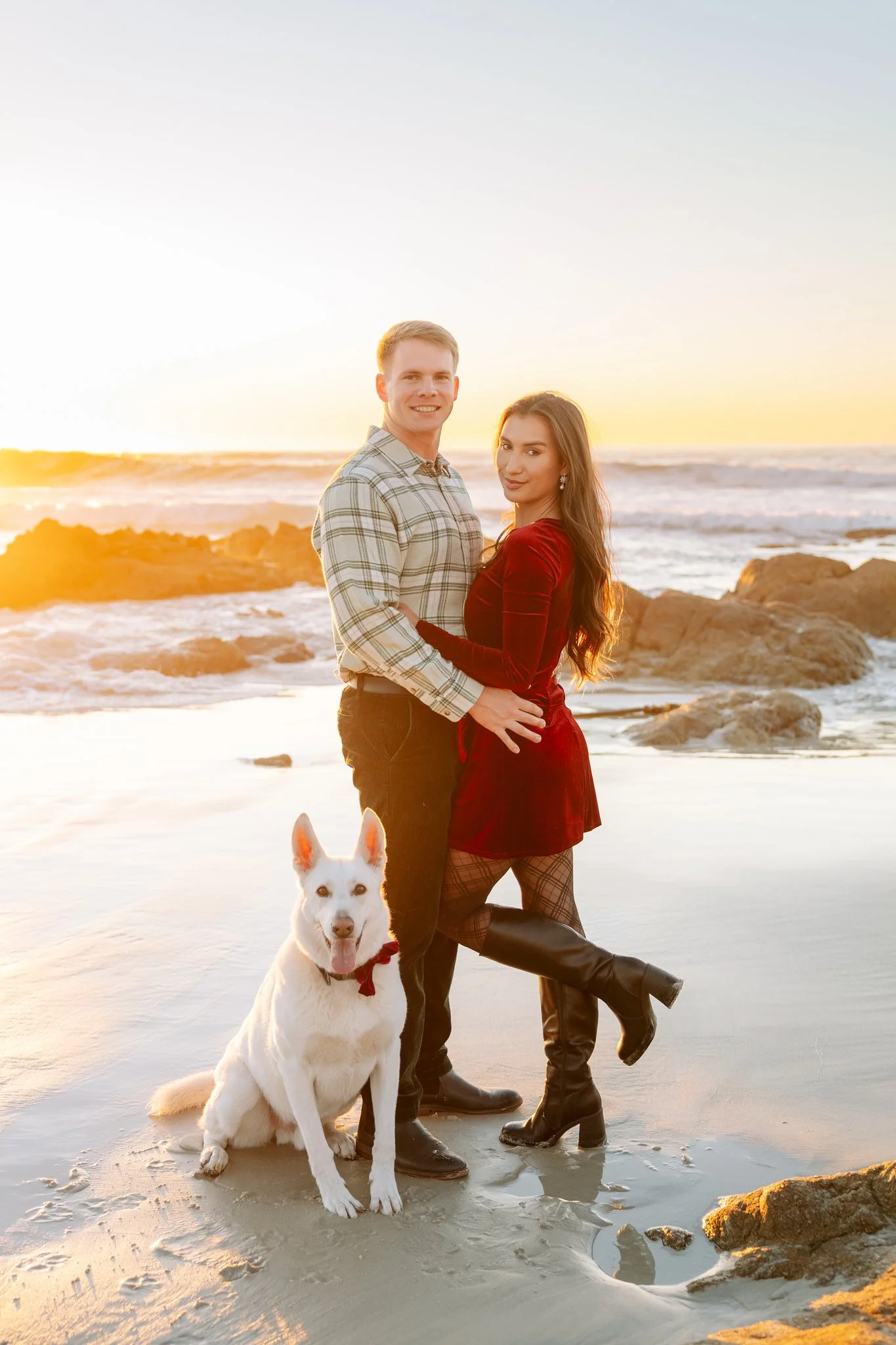 Husband, wife, and dog stand on the beach at sunset for a Christmas portrait.