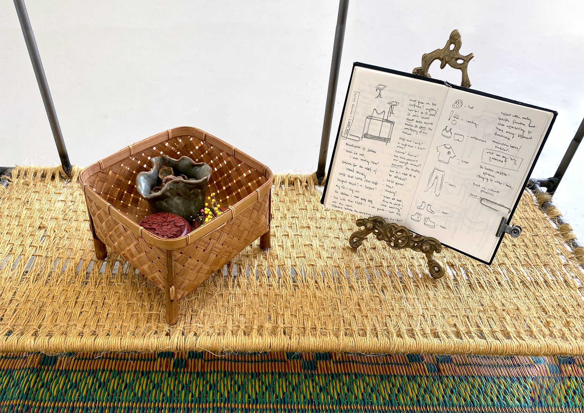 Atop a colorful, woven mat rests a two tiered metal stand. The second tier at ankle height holds a basket with a small ceramic vessel, a pin cushion, and a red box. Next to the basket is a brass picture stand holding an open sketchbook.