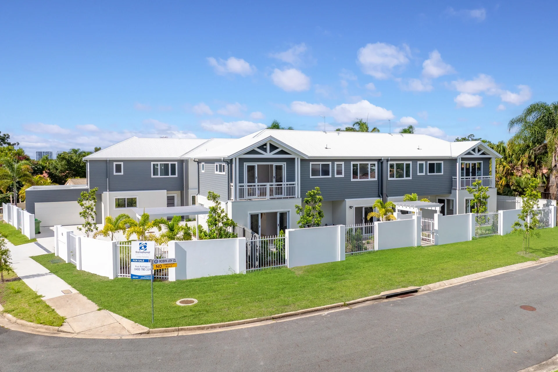 Large modern gray-and-white suburban home with balconies, fenced tropical landscaping, and a real estate for-sale sign