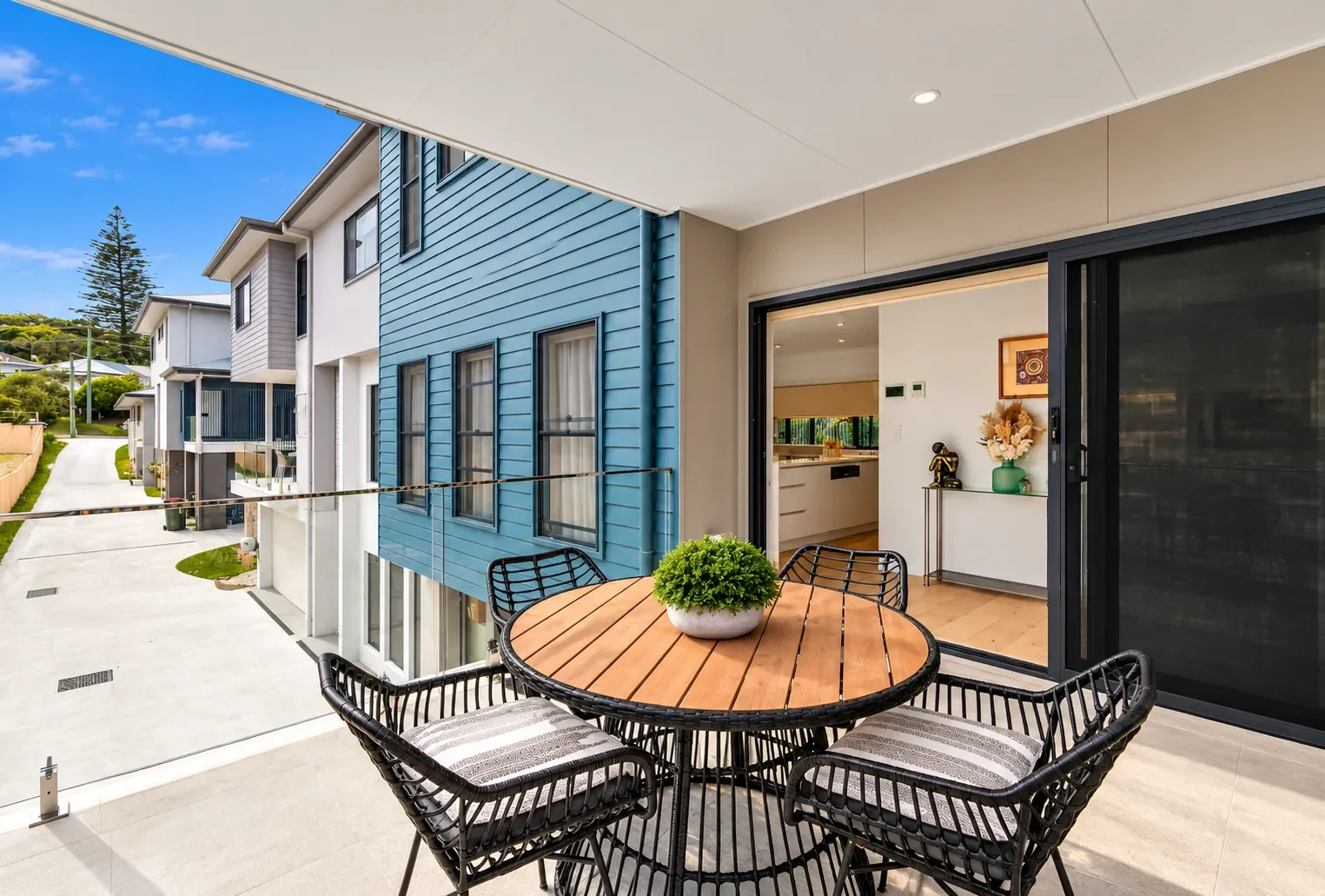 Modern outdoor patio with a round wooden table, black wicker chairs, and open doors to a bright contemporary home