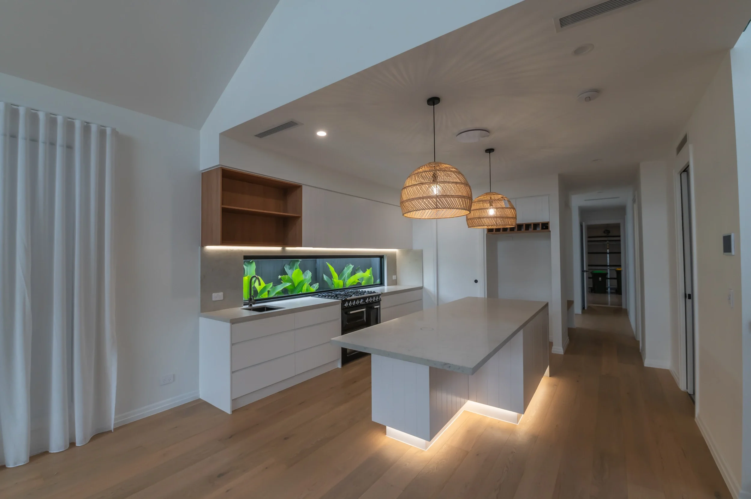 Modern white kitchen with large island, pendant lights, gas range, wood cabinetry, and indoor garden window