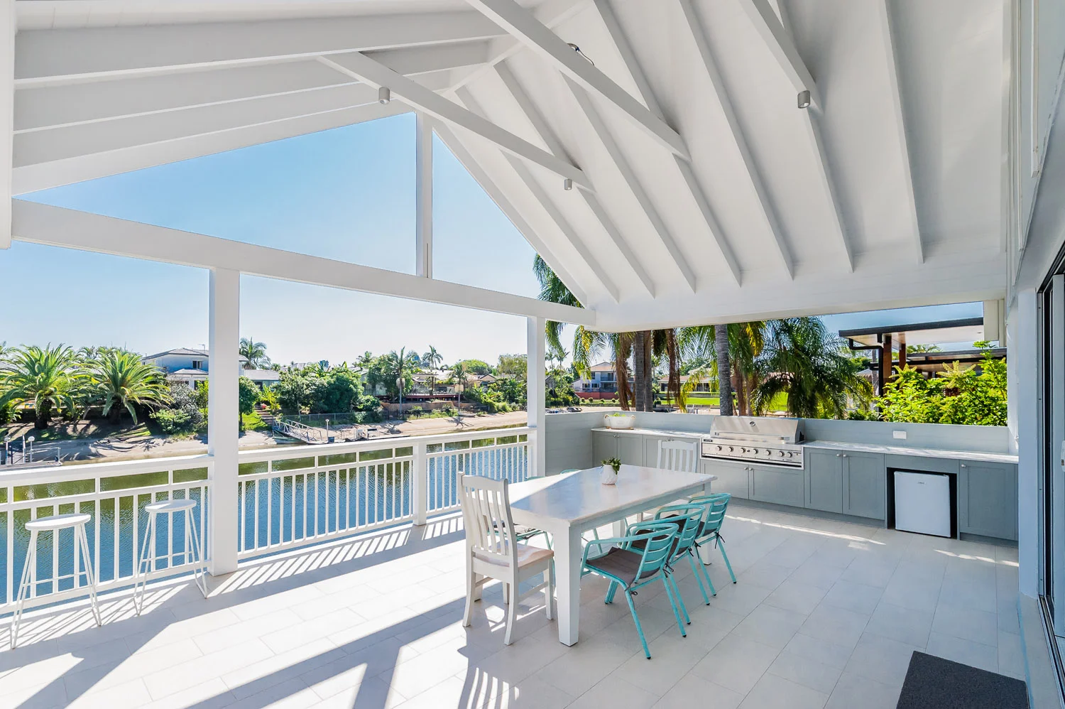 Covered waterfront patio with outdoor dining table, turquoise chairs, built-in grill, and canal view
