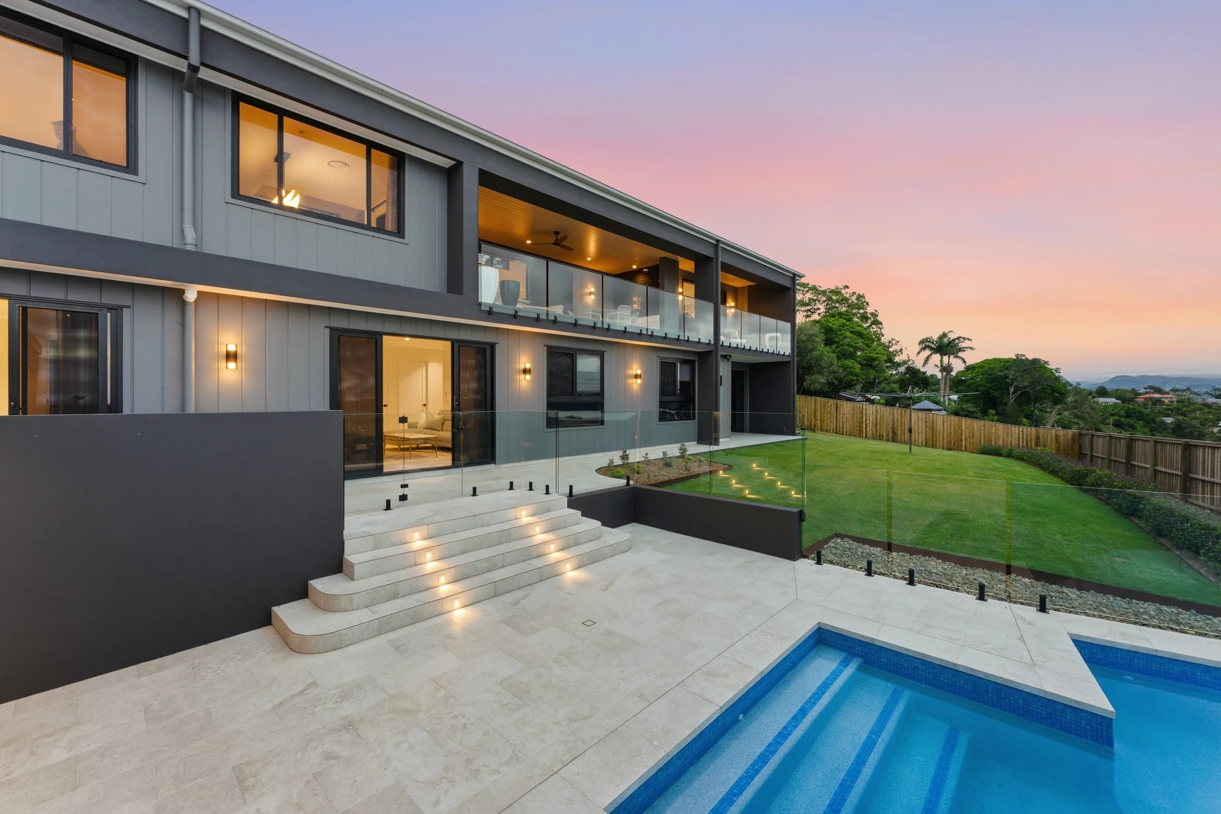 Modern two-story home with a glass-railed balcony, landscaped yard, outdoor pool, and mountain views at sunset