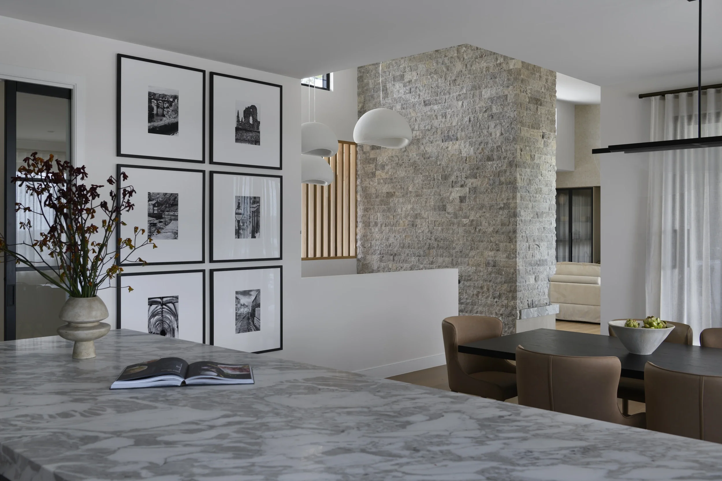 Modern open-plan home interior with marble kitchen island, stone accent wall, framed black-and-white artwork, pendant lights, dining table, and decorative vase
