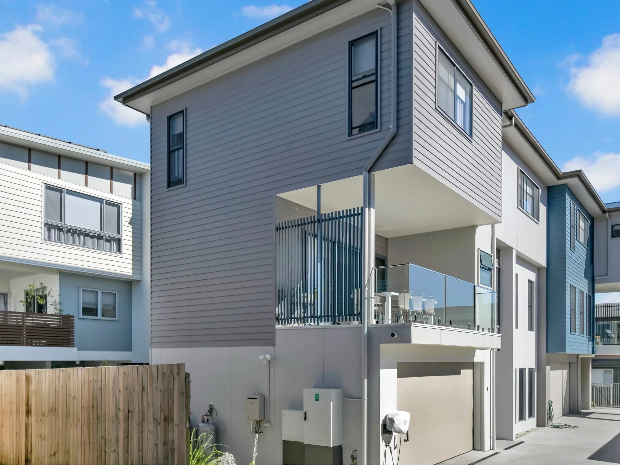 Modern multi-unit townhomes with gray siding, balconies, garages, and outdoor patios under a blue sky