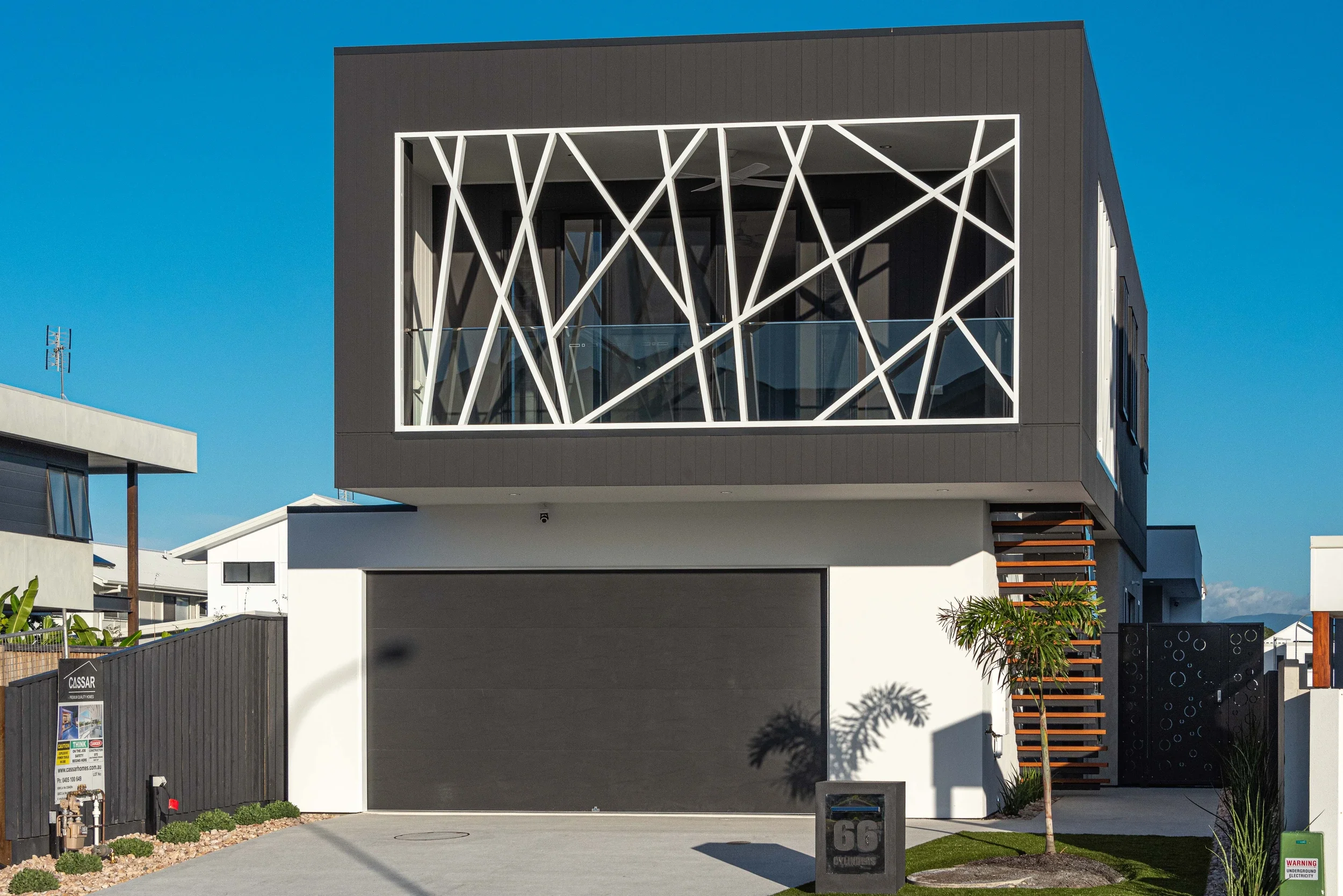 Modern two-story home with dark exterior, white geometric window framing, garage, balcony, tropical landscaping, and blue sky