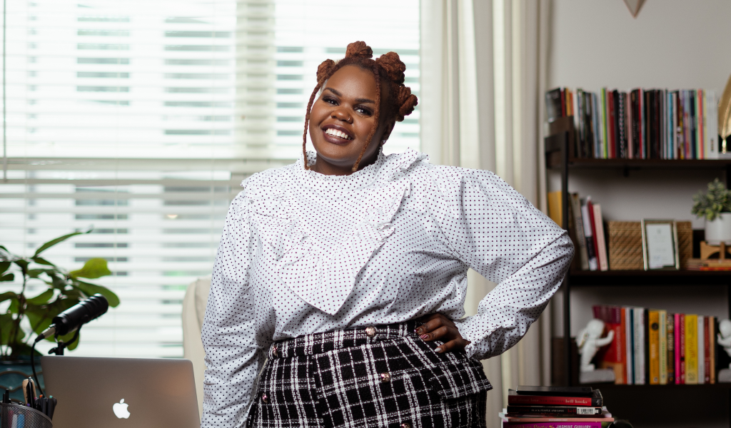 A smiling woman with braided hair in a bun, wearing a white polka dot blouse and plaid skirt, standing in a modern home office with a desk, laptop, and books.