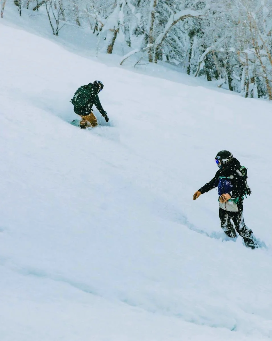 Two people dressed in winter gear, including helmets and goggles, are snowboarding and walking in deep snow on a snowy slope with snow-covered trees in the background.