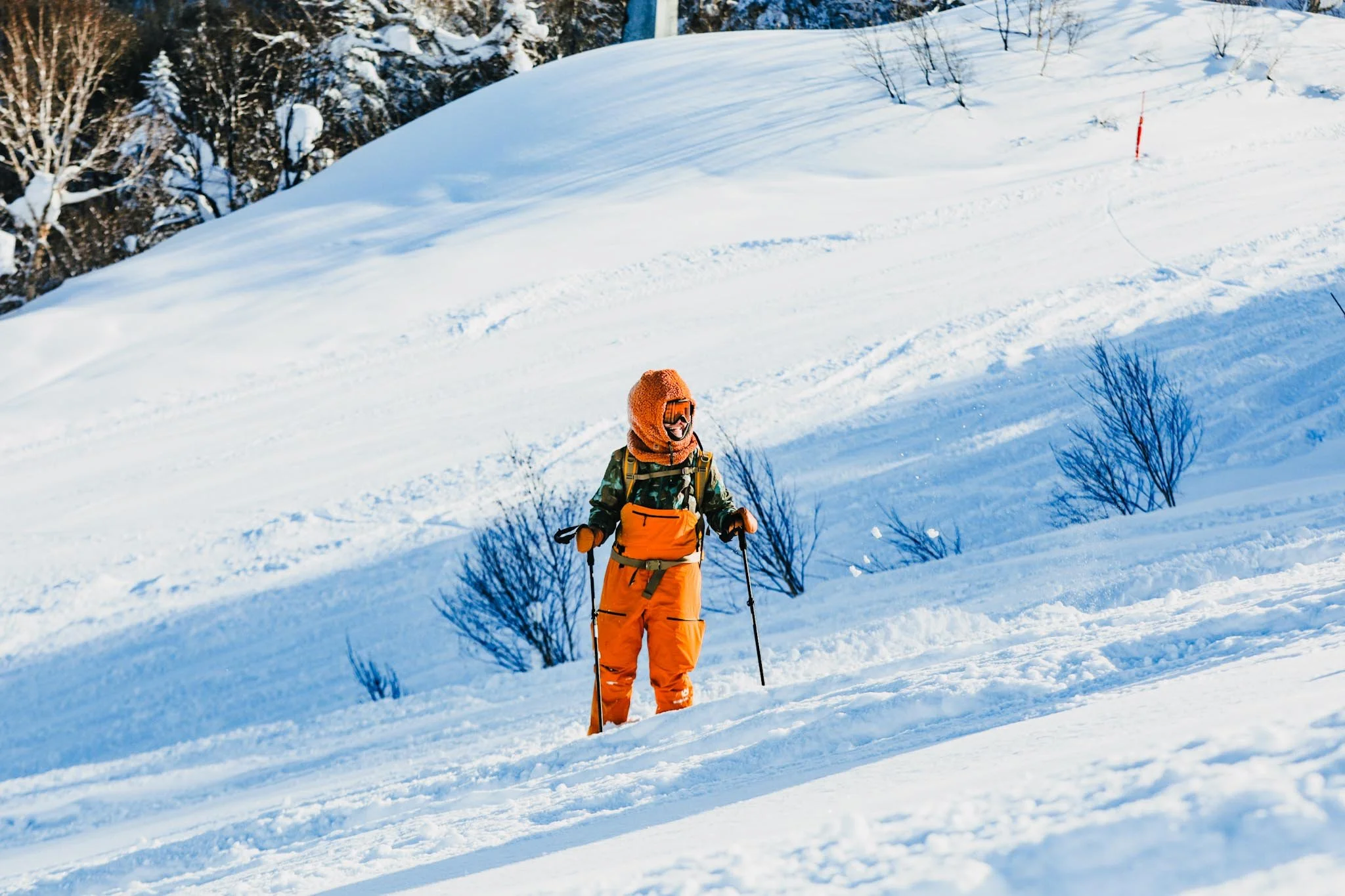 A person dressed in orange snow gear with a hoodie and sunglasses, carrying a backpack, skiing on a snowy mountain slope with trees and a red marker in the background.