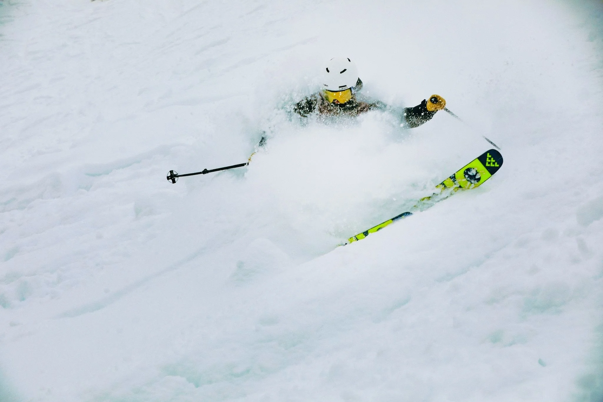 A skier in a yellow and black suit has fallen in deep snow while skiing down a slope and is partially buried in snow with ski poles and skis visible.