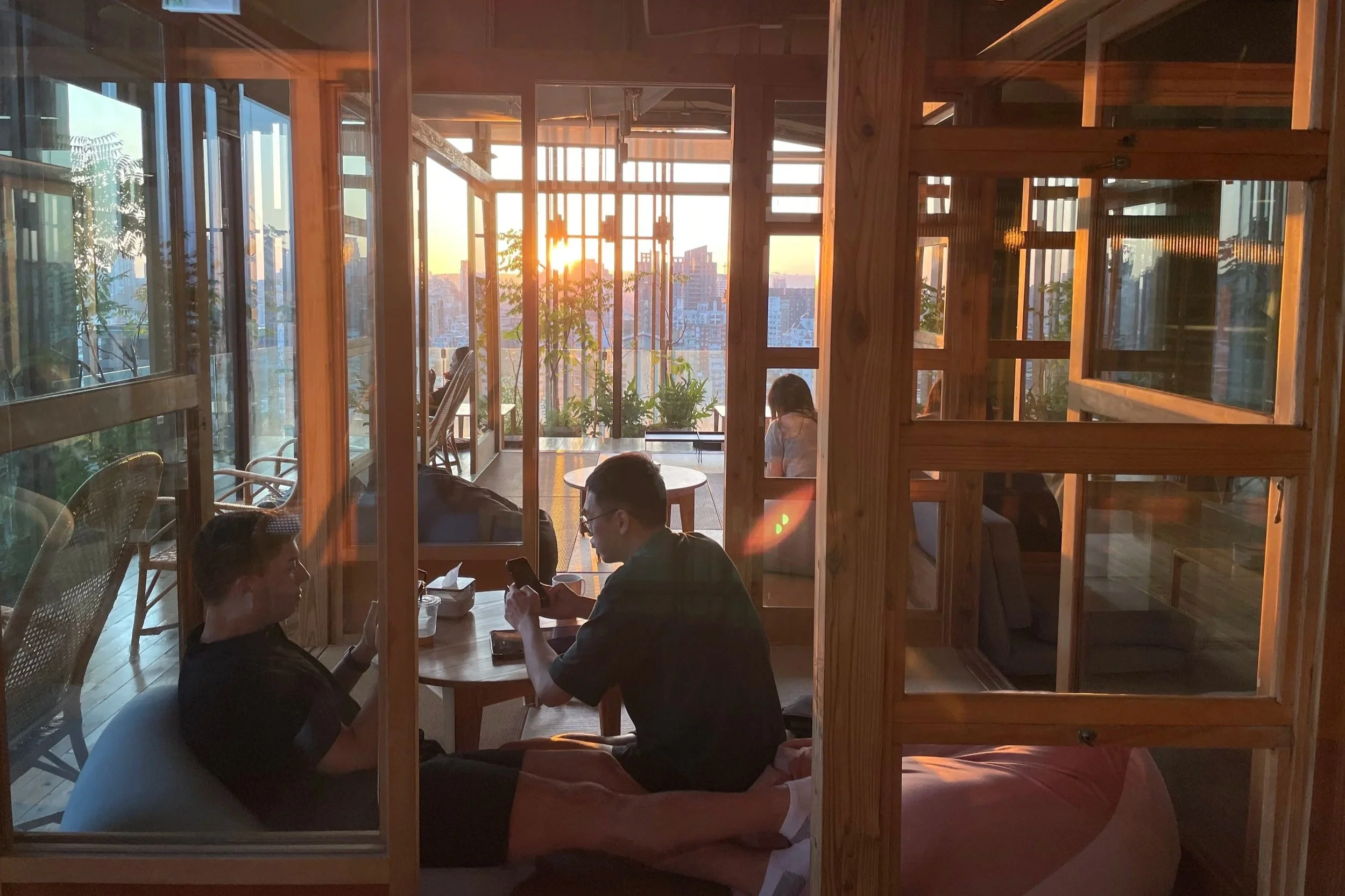 Inside a cozy café with large windows, warm sunlight streaming in, wooden furniture, and people sitting at tables, one person using a phone, with city buildings visible in the background.