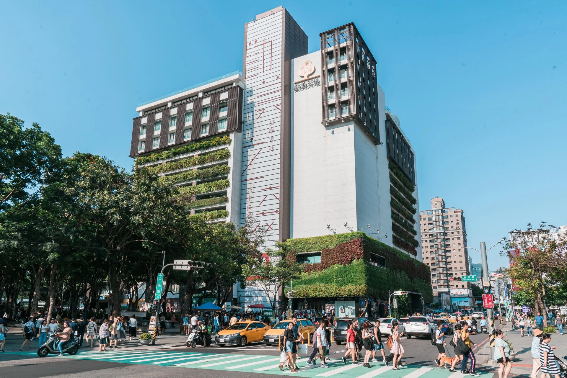 Busy city crosswalk with pedestrians, taxis, and scooters under a tall modern building with greenery, street signs, and trees, under a clear blue sky.