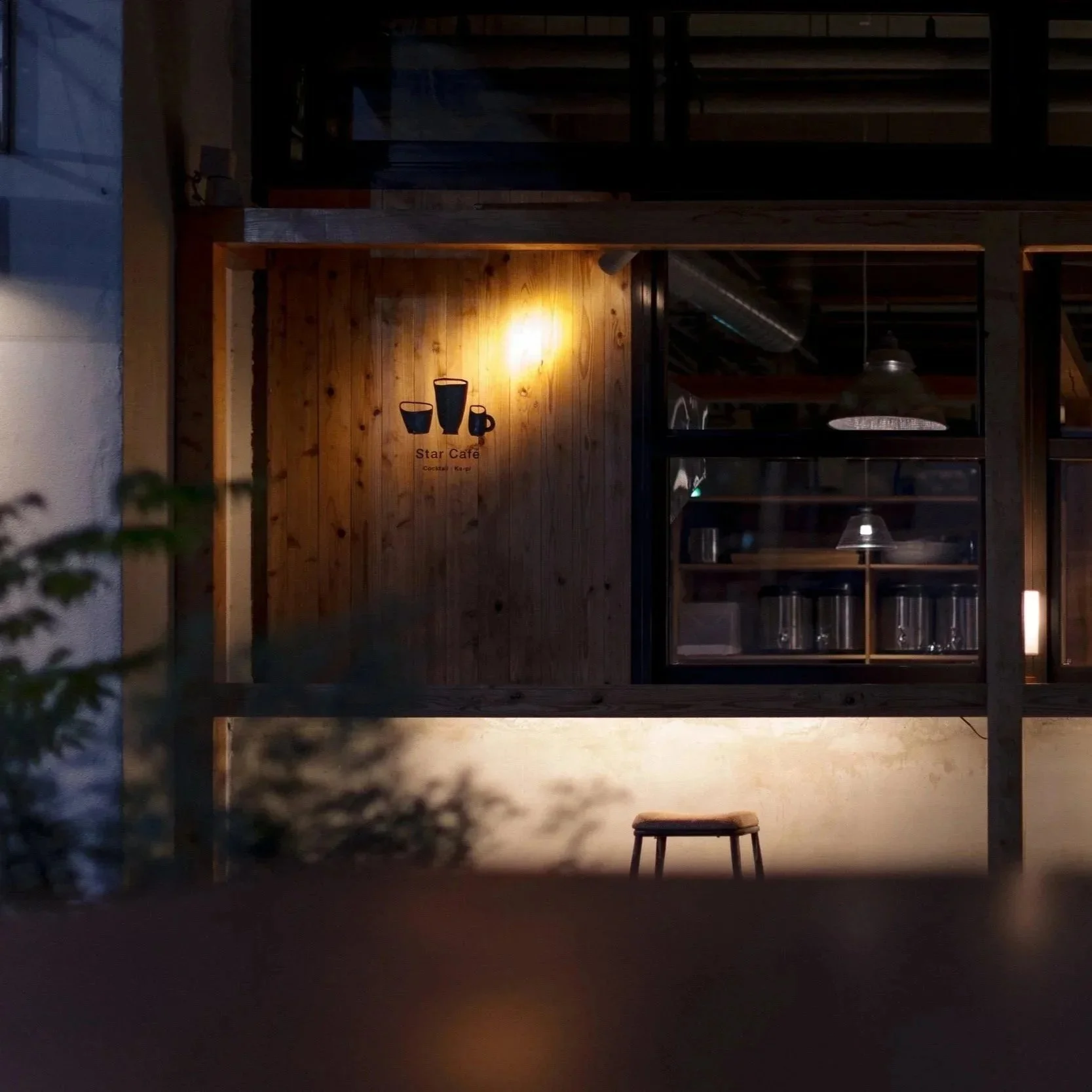 Nighttime view of a cafe exterior with a wooden wall, illuminated sign featuring black cups, and a single stool outside. The scene is dimly lit, creating a cozy ambiance.