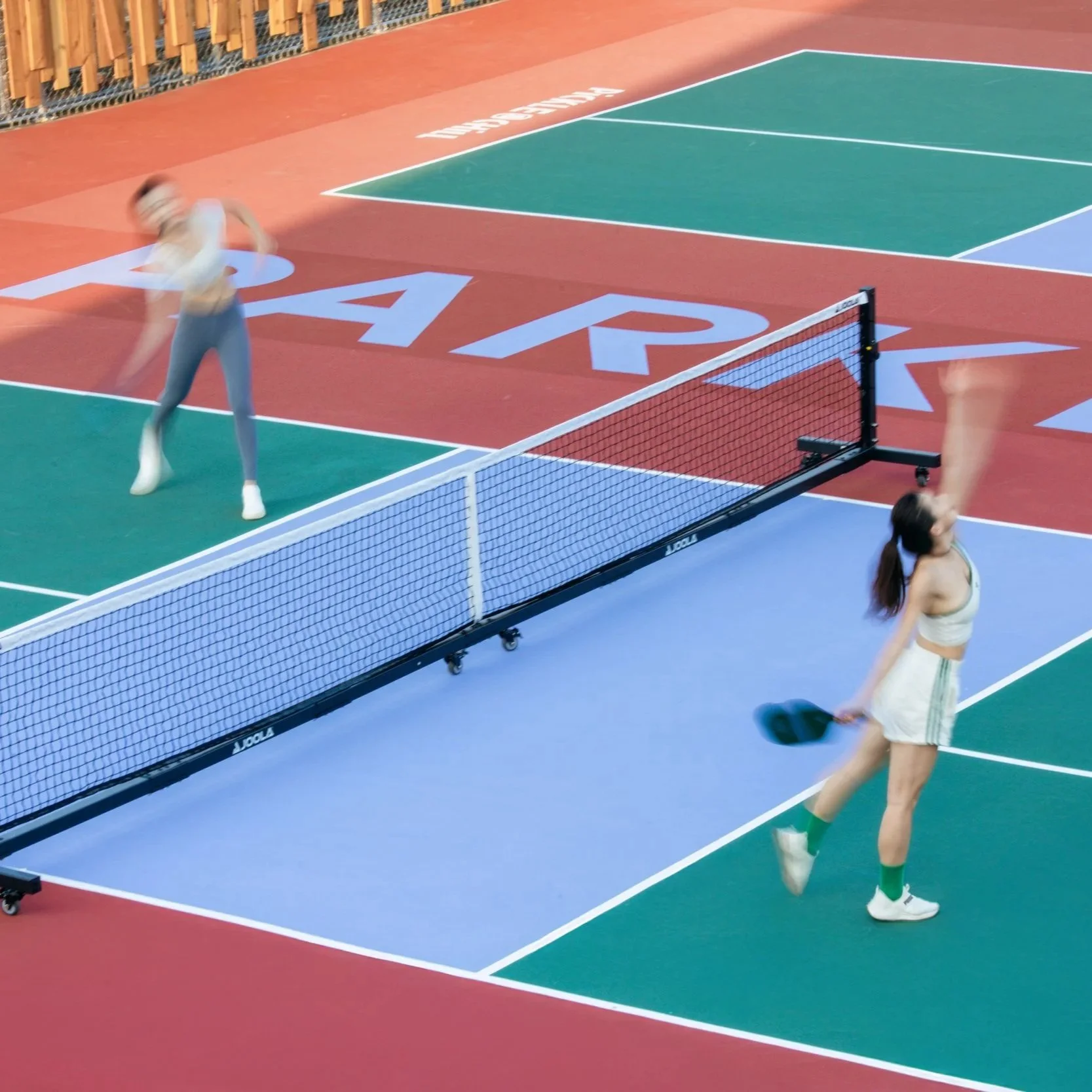 Blurry image of two women playing pickleball on an outdoor court with colorful surface and net, with a fence in the background.