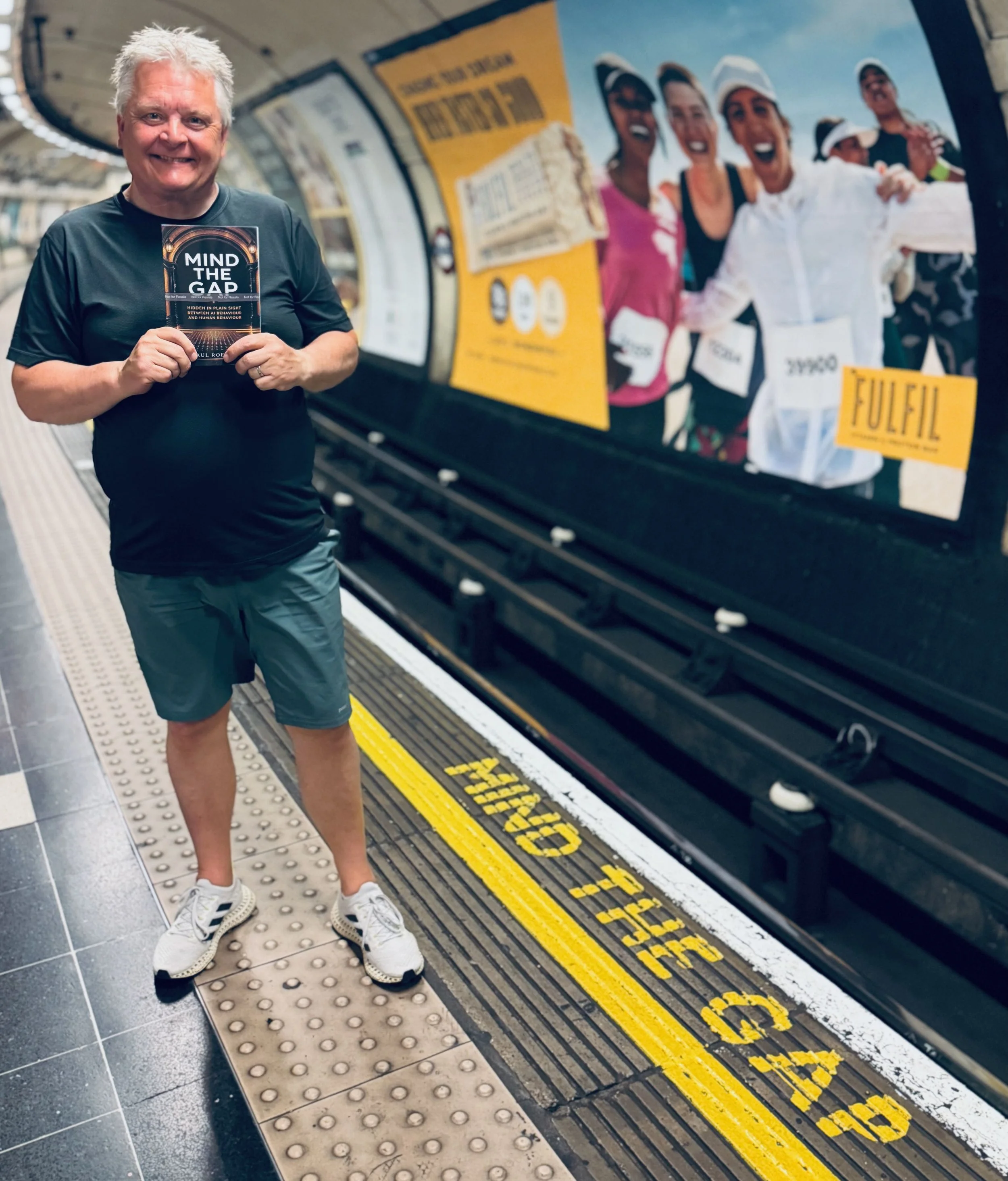 Paul Roebuck on a London Underground platform holding his book beside the painted words Mind the Gap