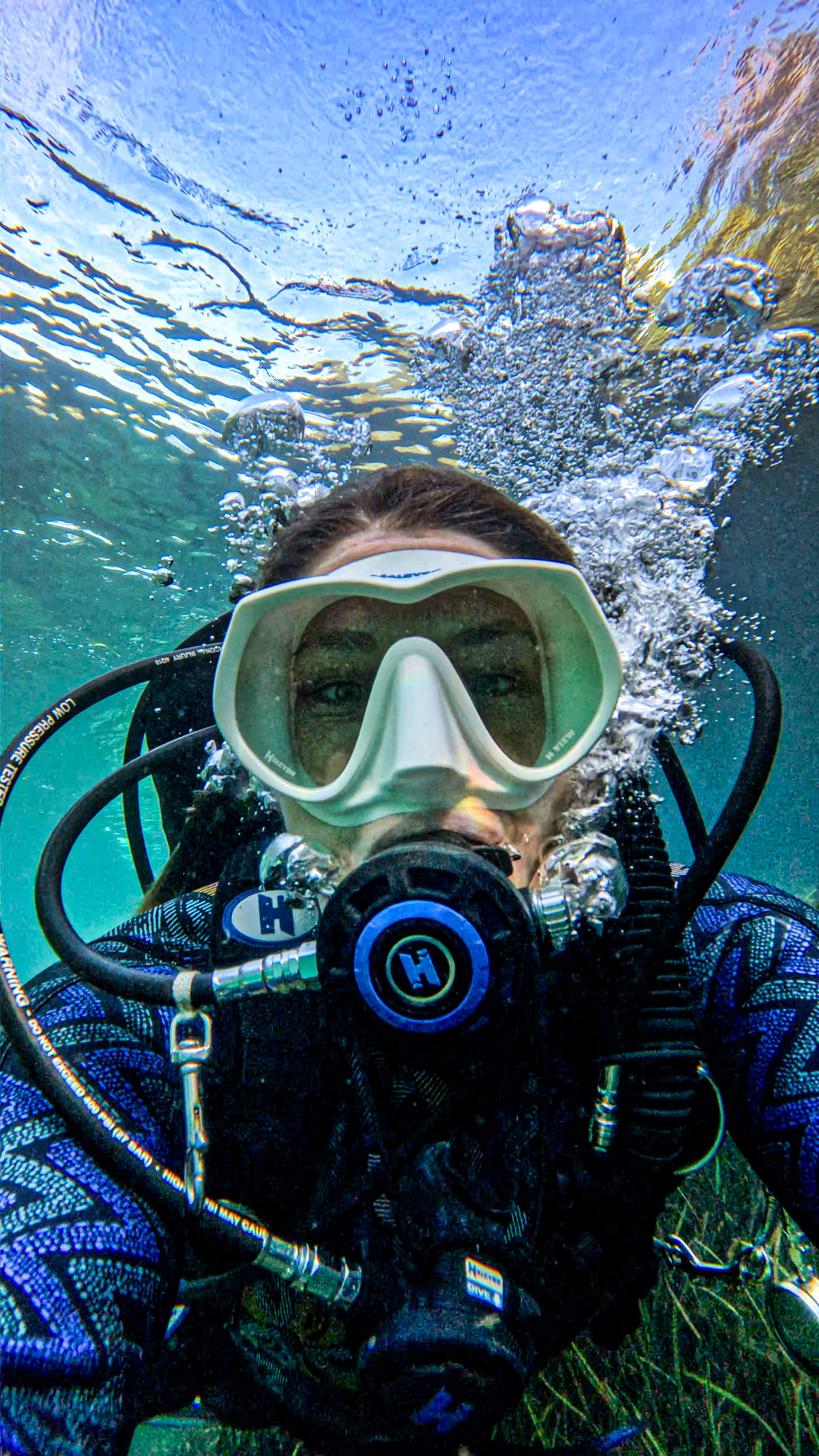 A person scuba diving underwater with a diving mask and breathing apparatus, with bubbles ascending toward the surface and coral visible at the bottom.