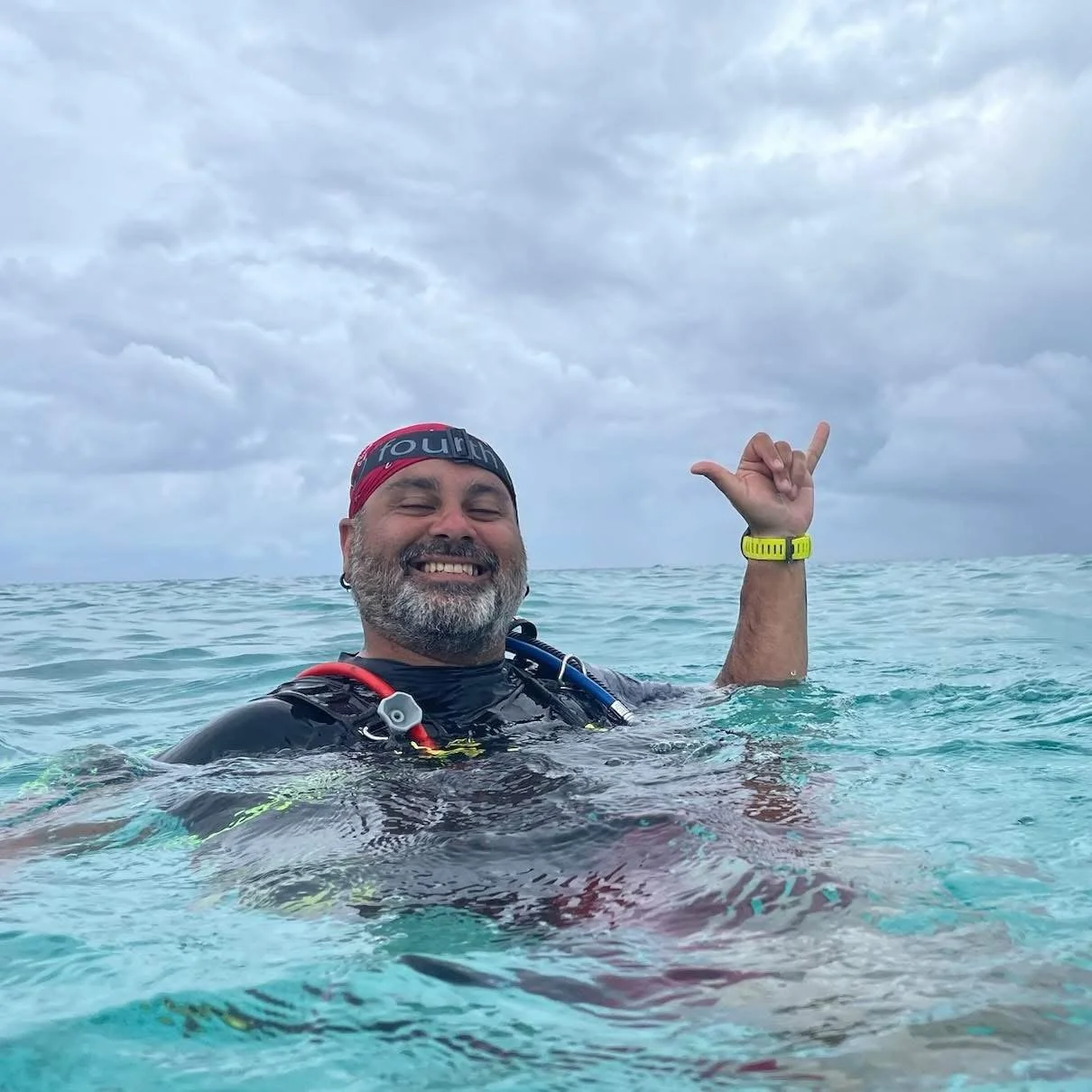 A man smiling in the ocean, making a shaka sign with his right hand, wearing a black wetsuit, a red and black swim cap, and a yellow wristwatch.