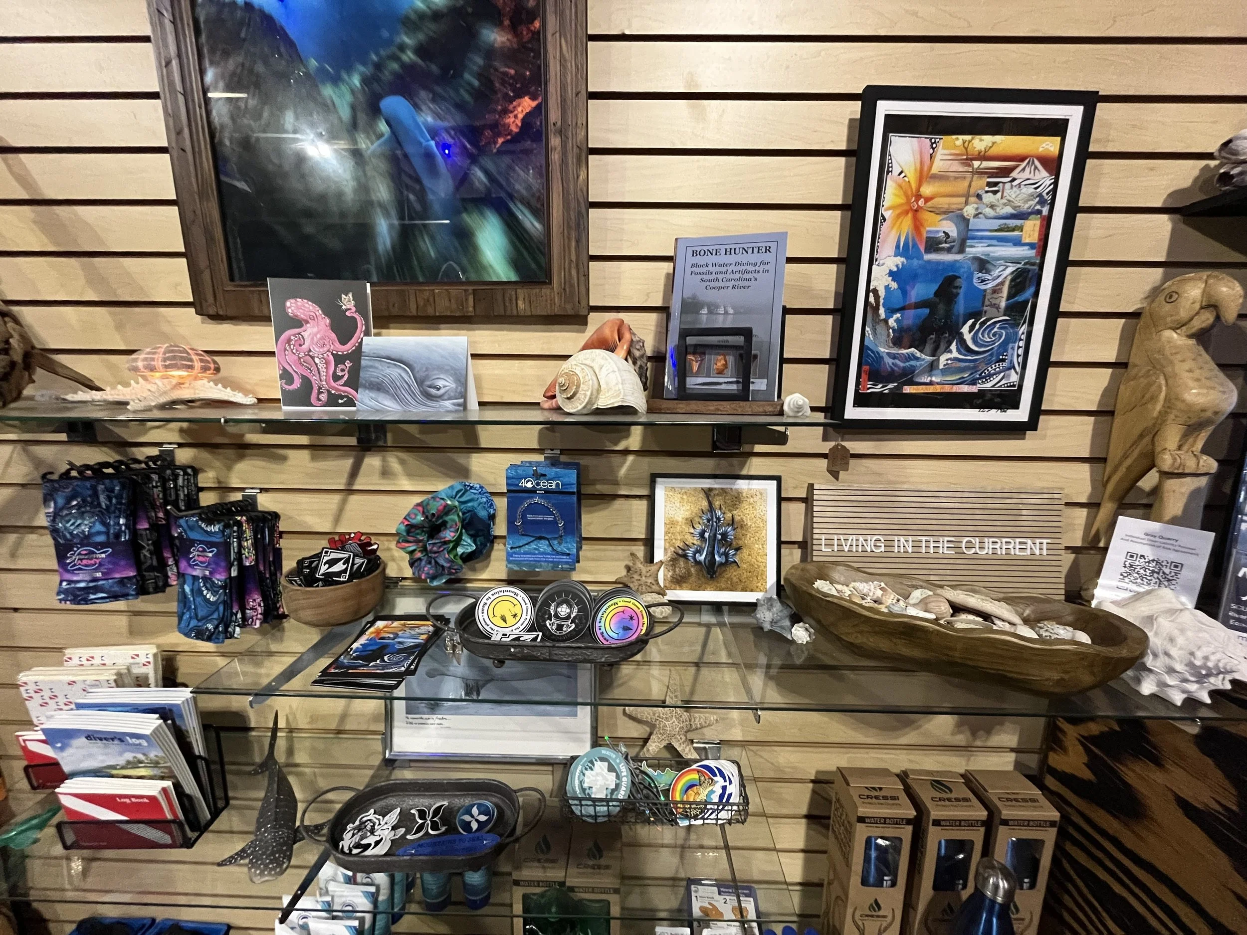 A display of ocean and nature-themed art, jewelry, and souvenirs on a glass shelf against a wooden slat wall.
