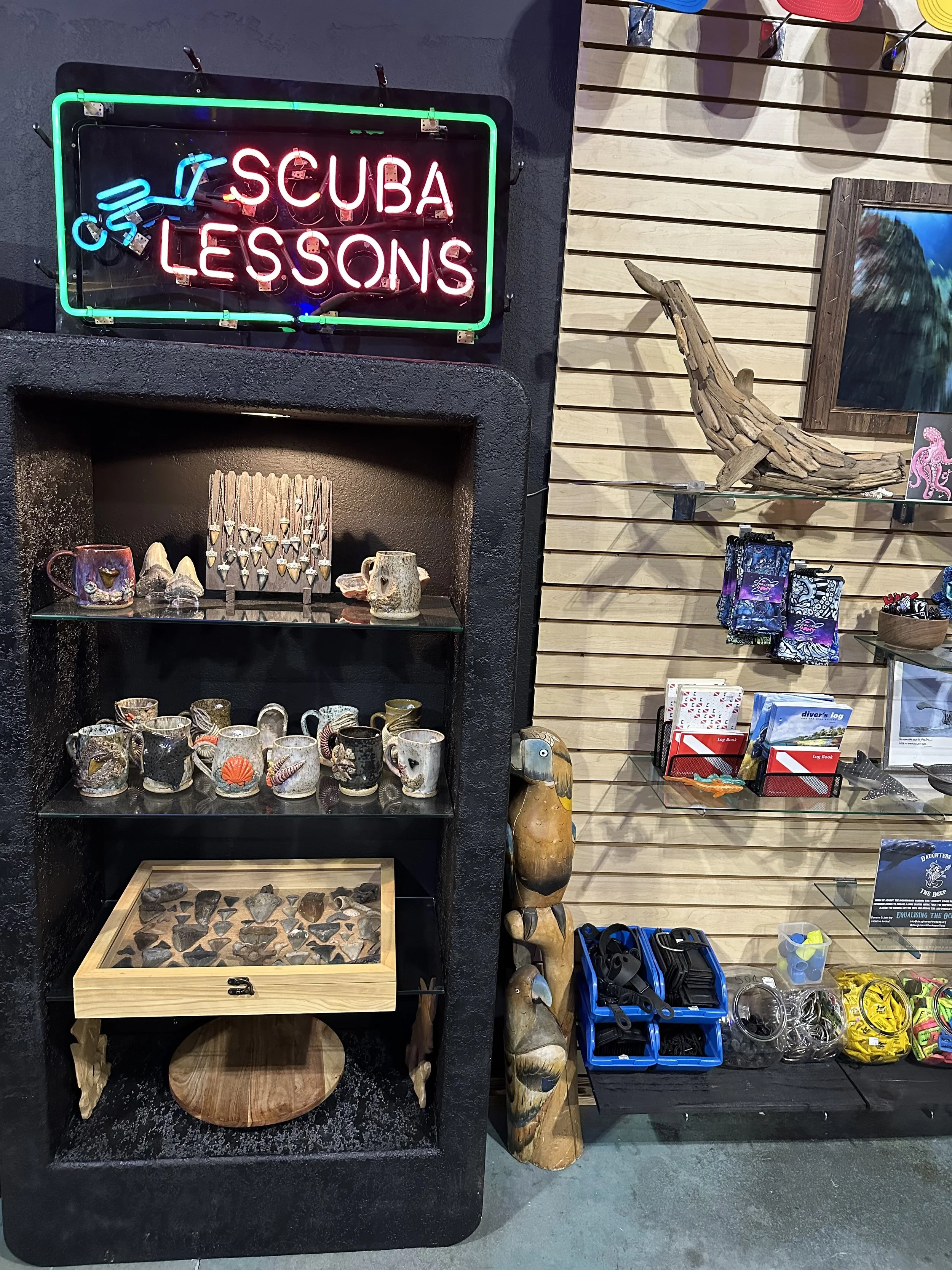 A display of souvenirs and decorative items on shelves, with a sign that says 'SCUBA LESSONS' in neon lights, including mugs, rocks, carved wooden totem, and other decorative pieces.