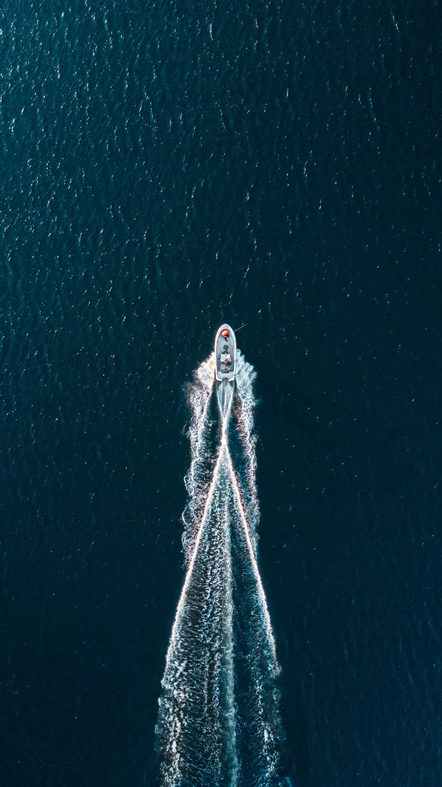 An aerial view of a boat moving through deep blue water, creating white wake patterns behind it.