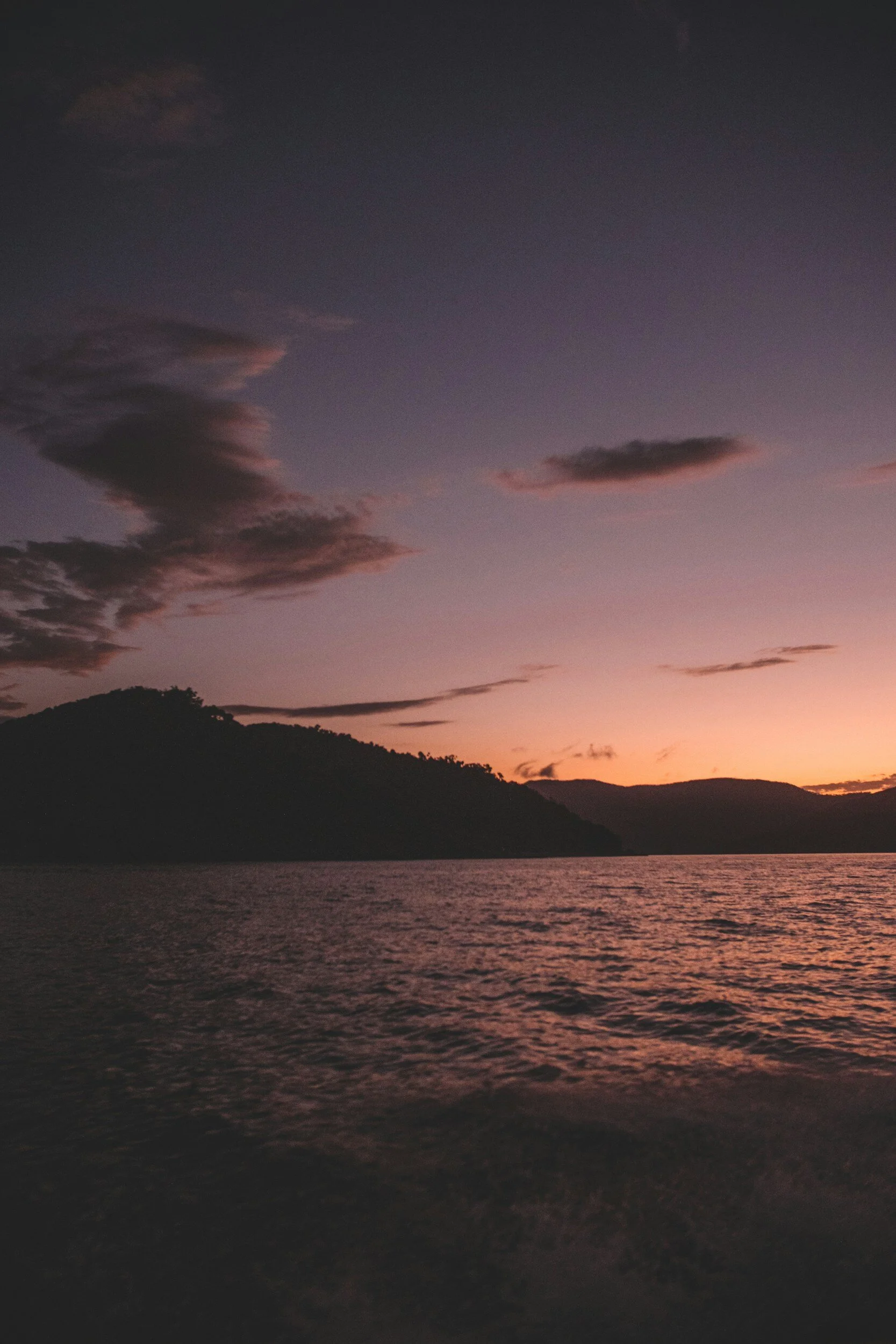 A tranquil scene of a lake during sunset with mountains in the background, colorful sky with scattered clouds, and gentle ripples on the water.