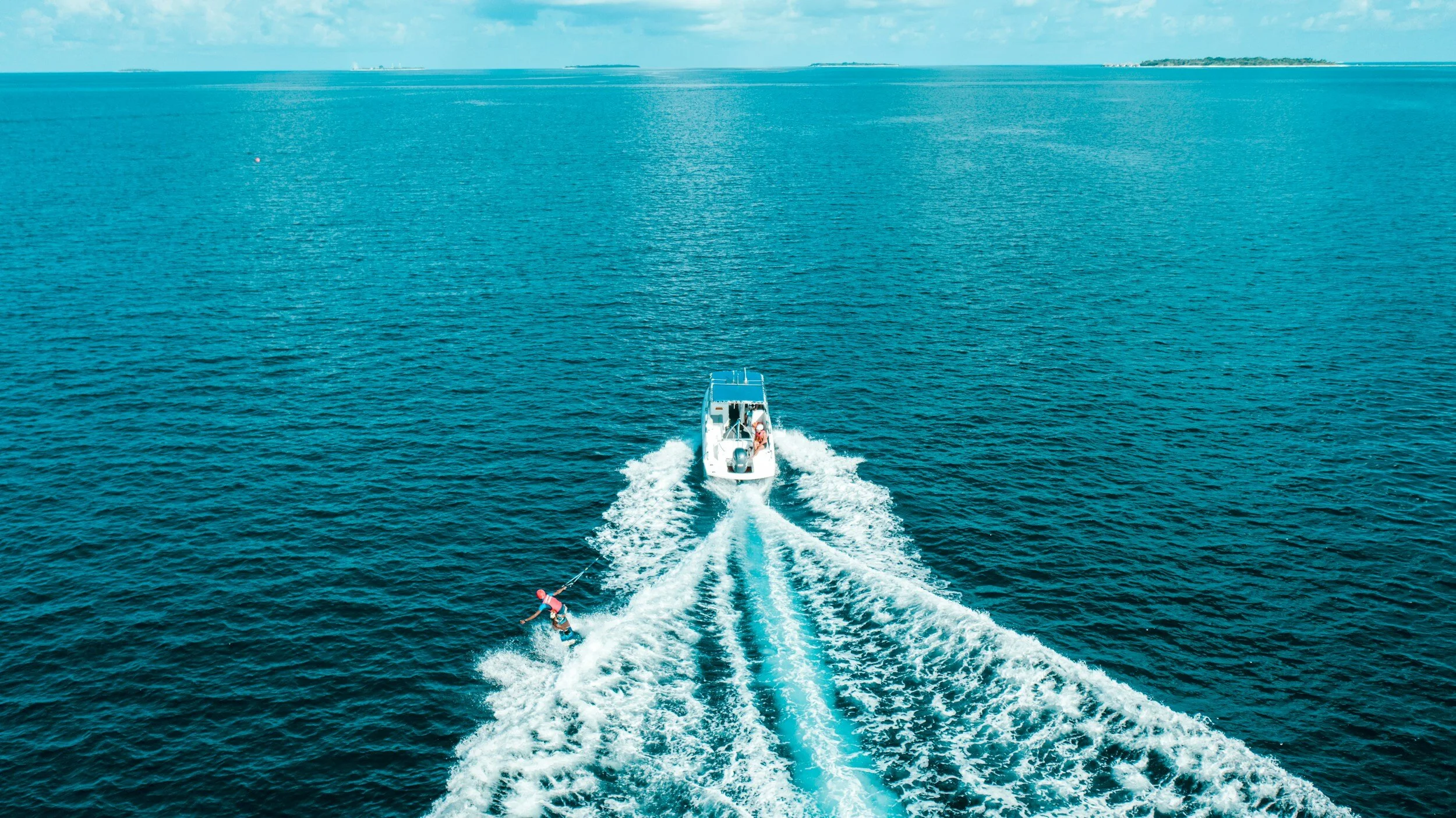 A boat pulling a person on a wakeboard in the open ocean with a distant shoreline in the background