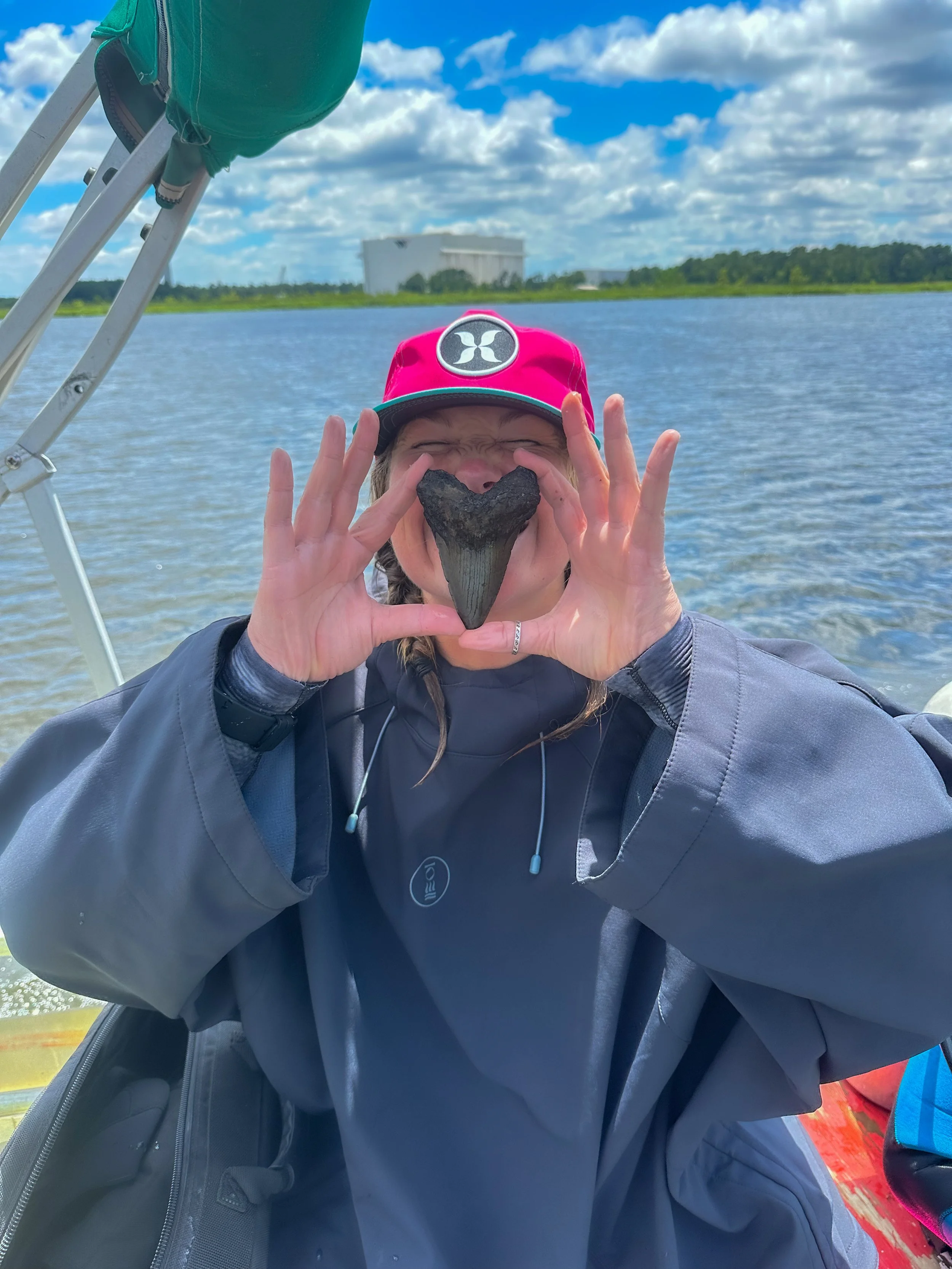 Person wearing a pink cap and gray jacket holding a large heart-shaped fish head in front of their face on a boat by a body of water with a cloudy sky and buildings in the background.