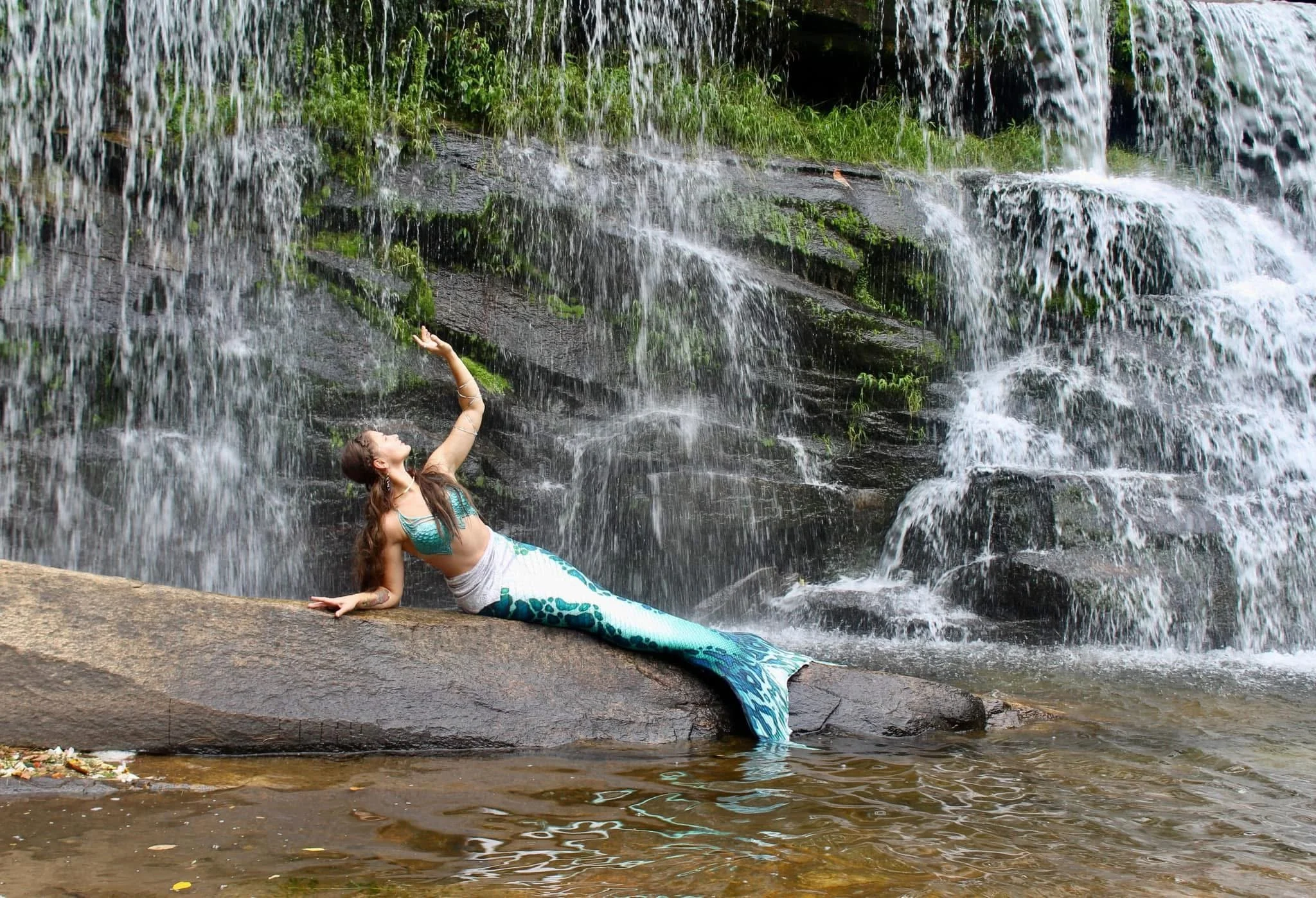 A woman dressed as a mermaid, with a turquoise and white tail and bikini top, lying on a large rock in front of a waterfall, reaching upward with one arm.