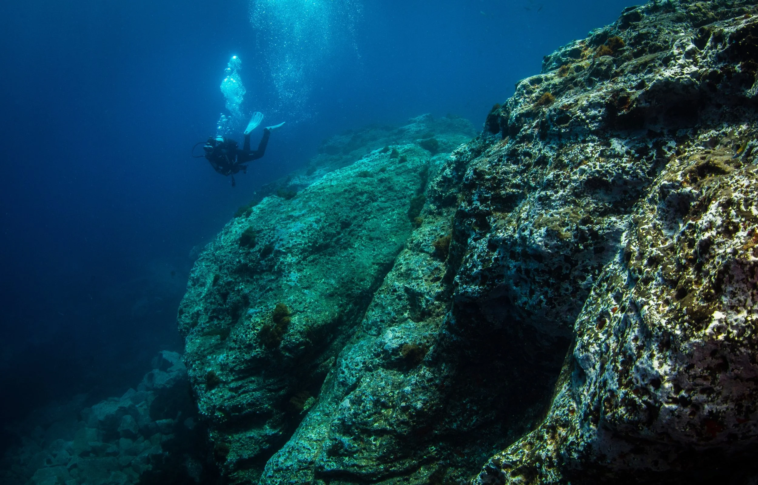 A scuba diver swimming near rock formations underwater in the ocean.