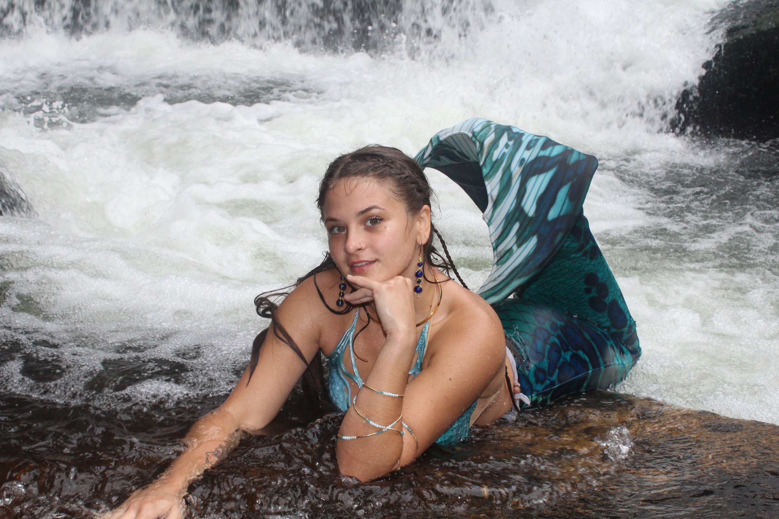 A woman with wet hair laying on a rock in a river, wearing a blue mermaid tail and swim top, with her chin resting on her hand, looking at the camera.