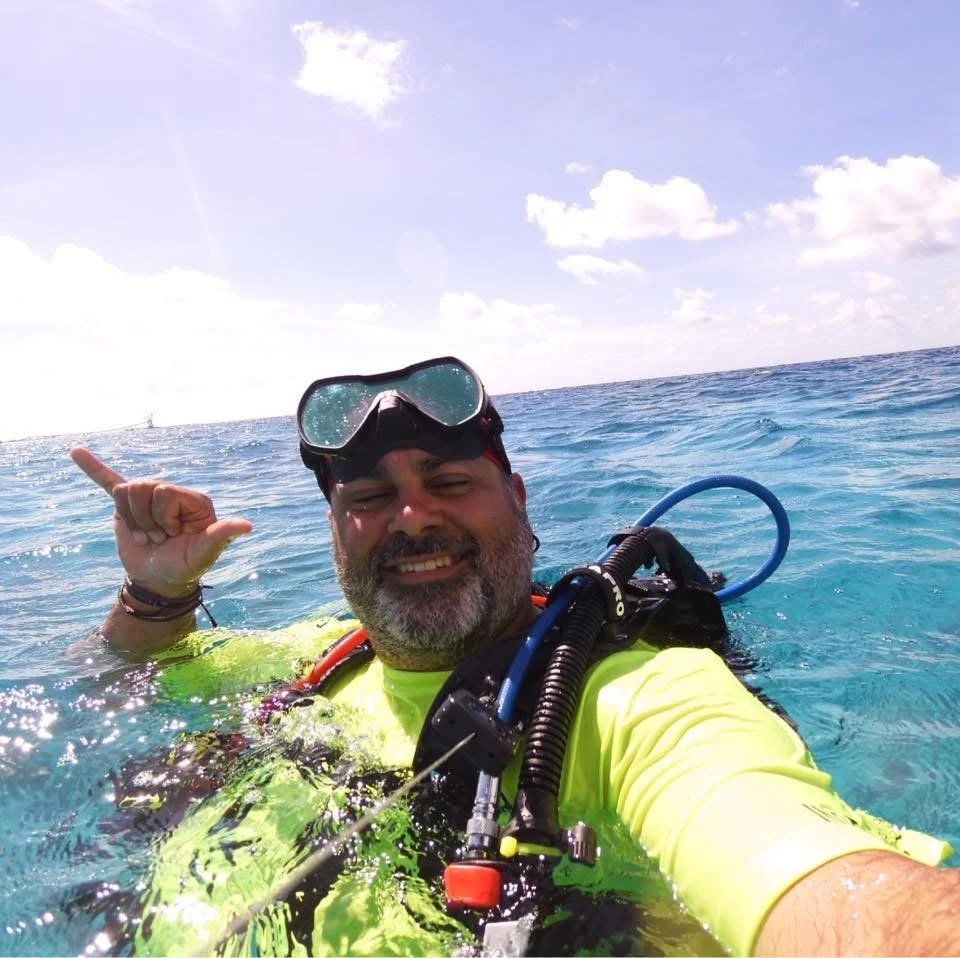 A man wearing scuba diving gear underwater on the ocean, smiling and making a hand gesture.