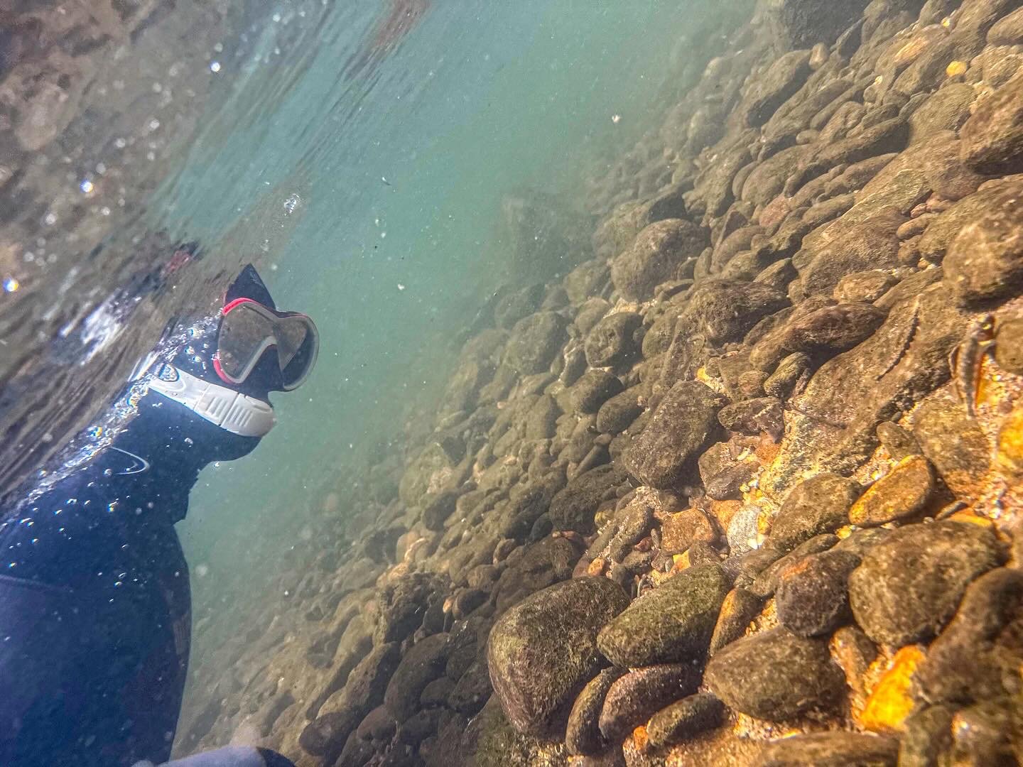 Underwater scene showing clear water, rocks on the riverbed, and a pair of slip-on shoes partially submerged in the water.