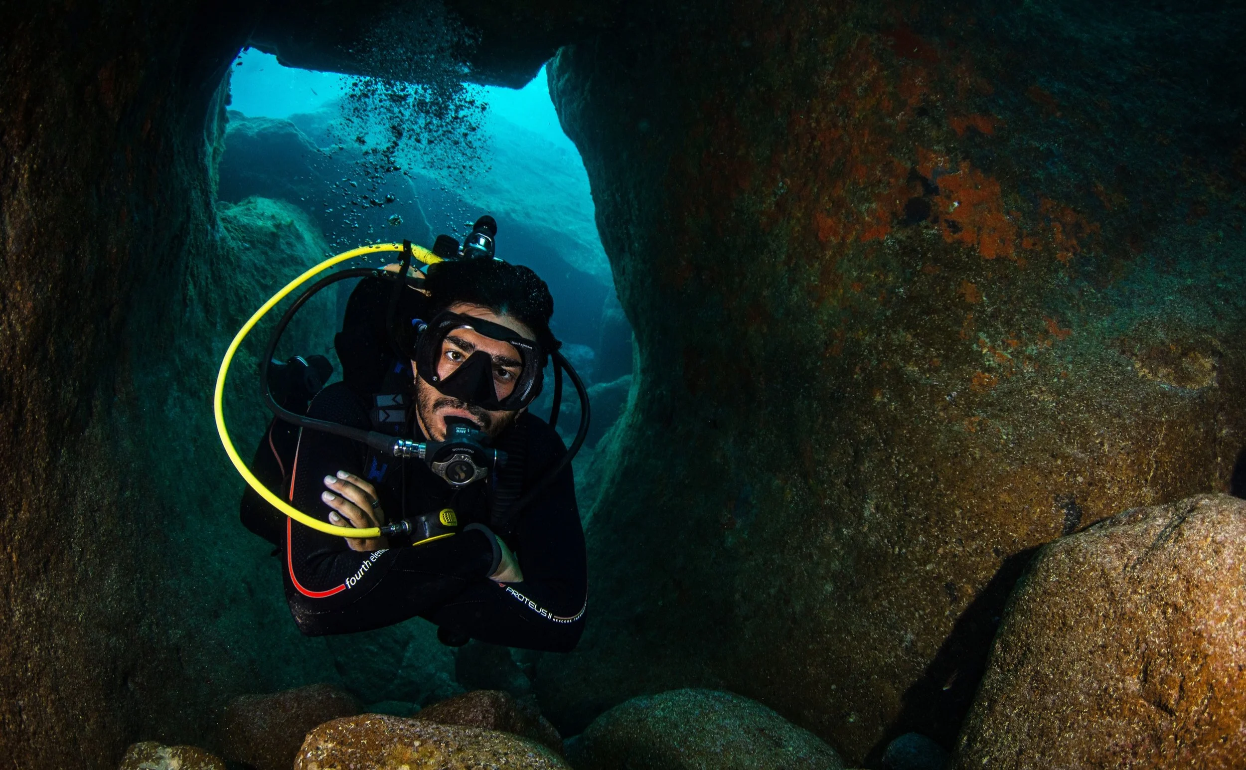 A male scuba diver with head and face mask, holding an underwater light, exploring through a rocky underwater cave or crevice, with blue water visible in the background.