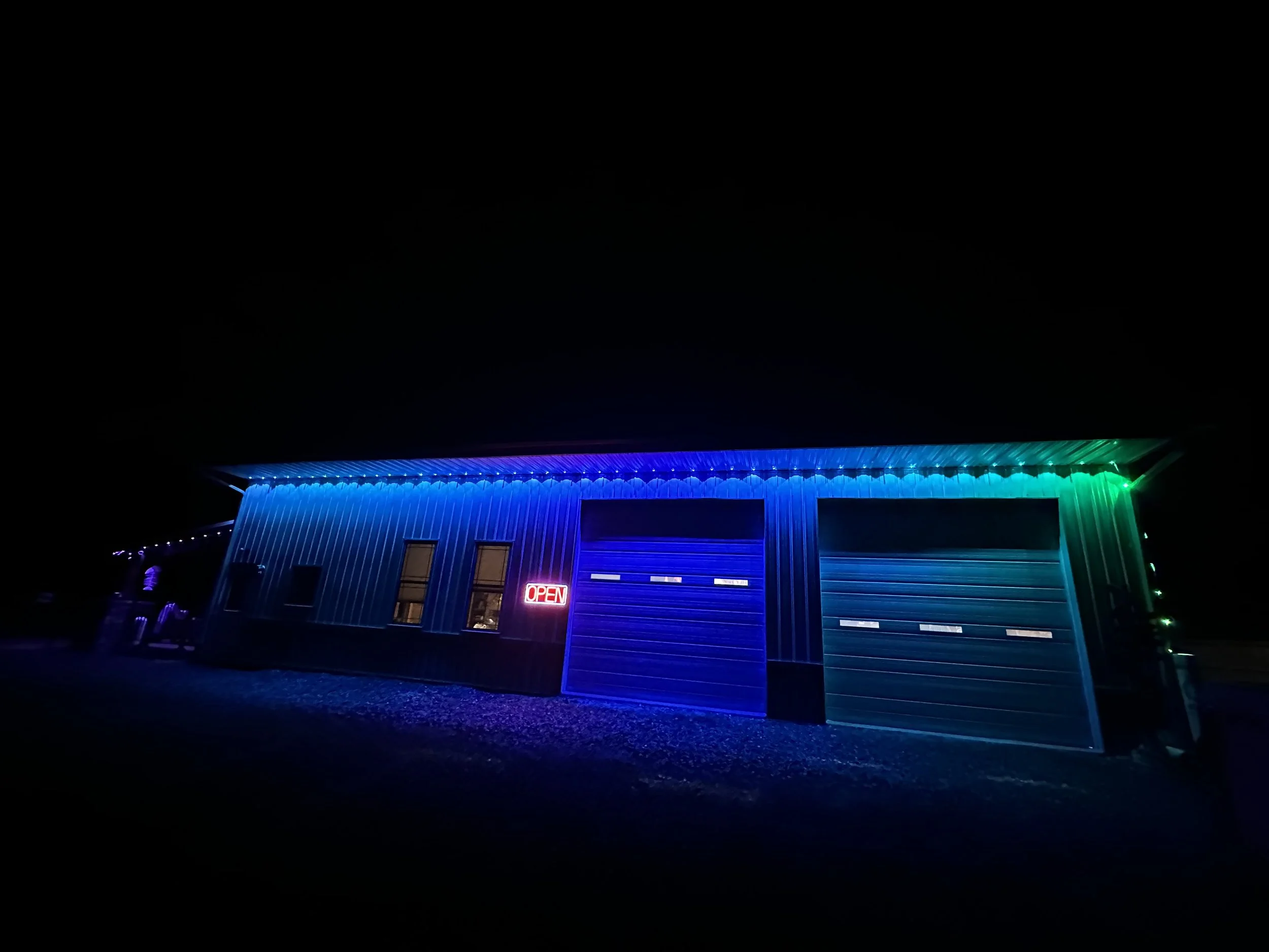 A building with blue and green LED lights along the roofline, a neon 'OPEN' sign, and two closed garage doors at night.
