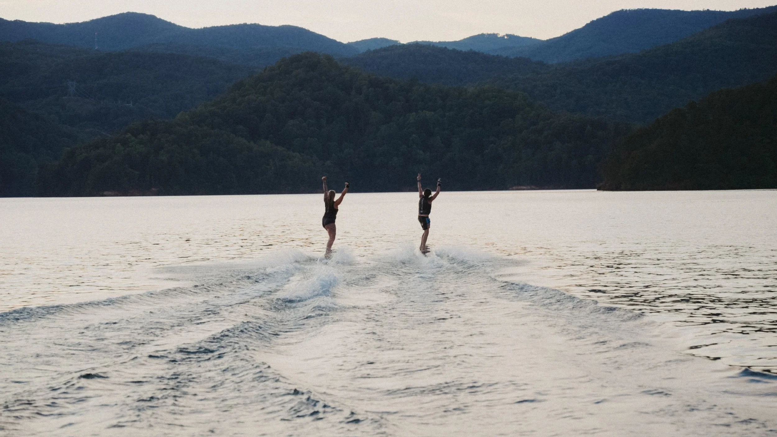 Two people wakeboarding on a large lake with mountains in the background during sunset.
