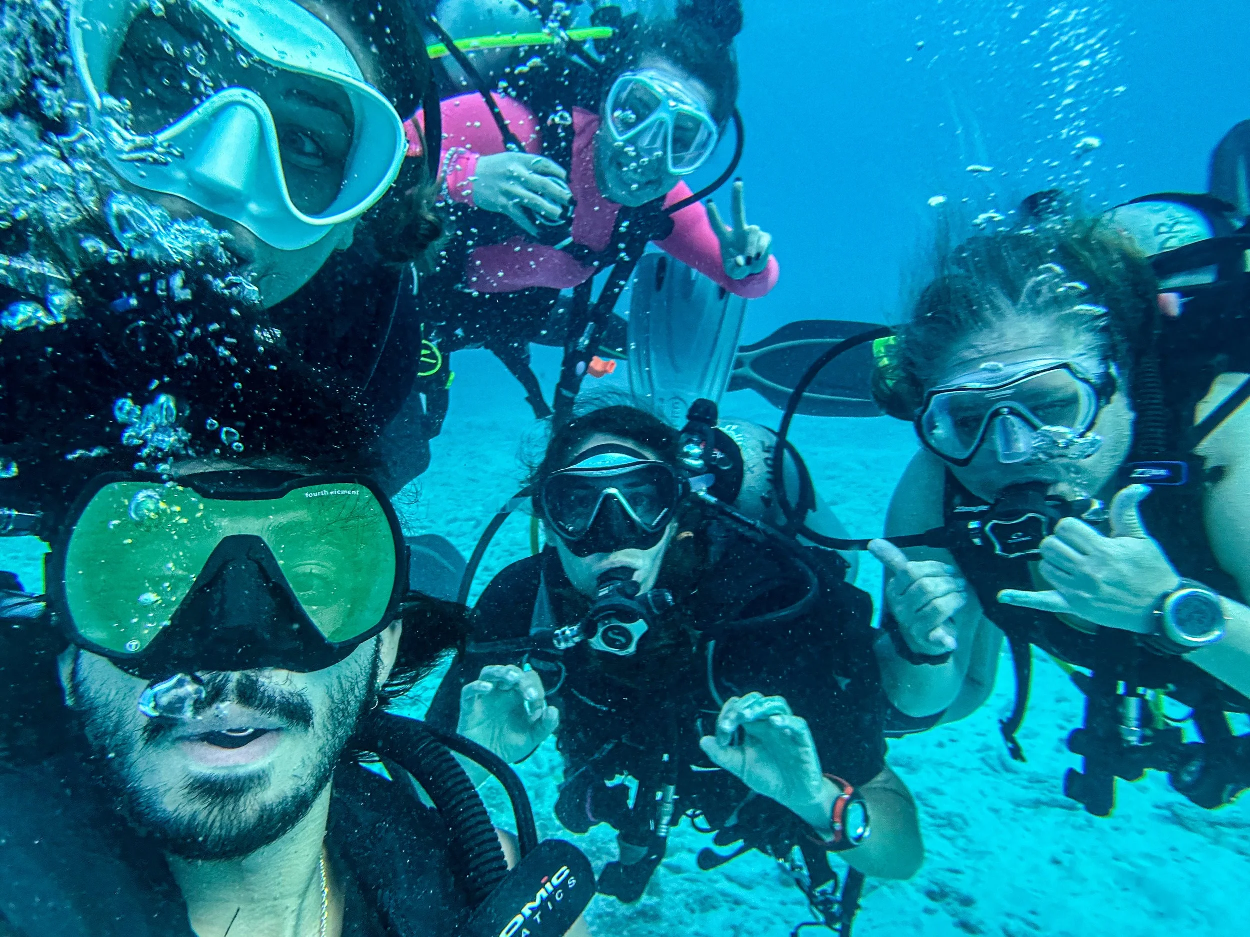 Four scuba divers posing underwater, making hand signs and taking a selfie.