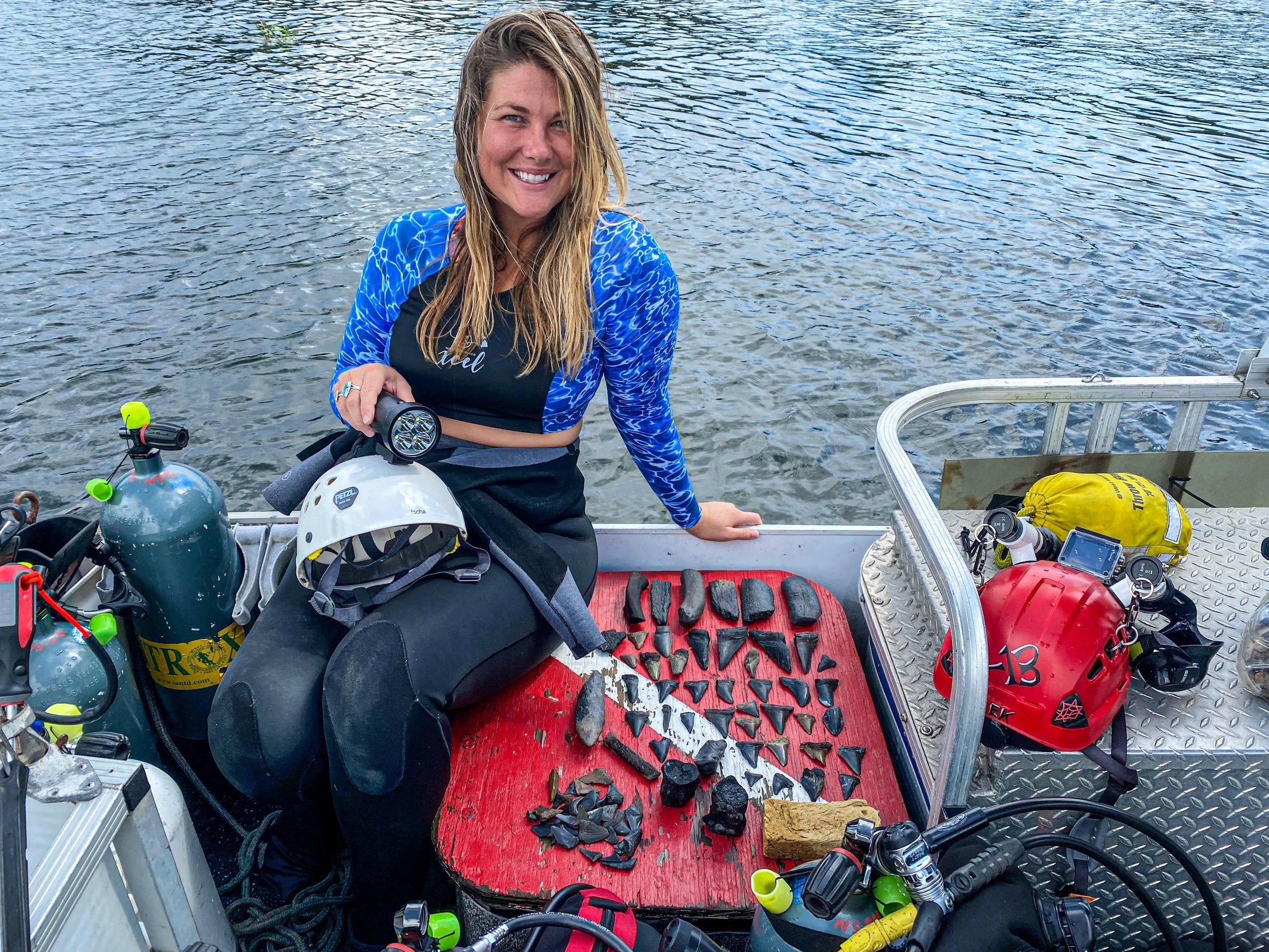 Woman smiling on a boat with rocks laid out on a red mat, surrounded by scuba diving equipment including tanks, helmet, and gauges.