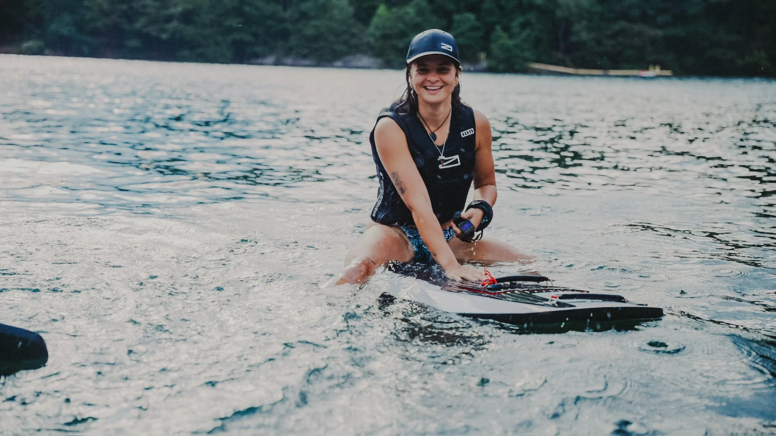 A person smiling and sitting on a wakeboard in a lake during daytime, with trees in the background.
