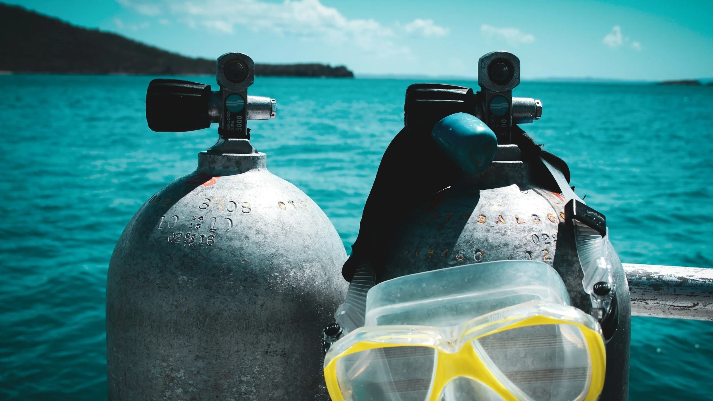 Two scuba diving tanks with a pair of yellow diving goggles resting in front, with a blue ocean and distant land in the background.