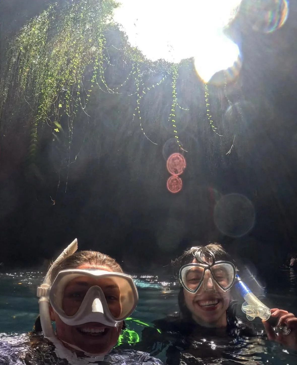 Two people wearing snorkeling masks smiling in a body of water, with sunlight coming from above and hanging plants or vines on the overhanging rocks.