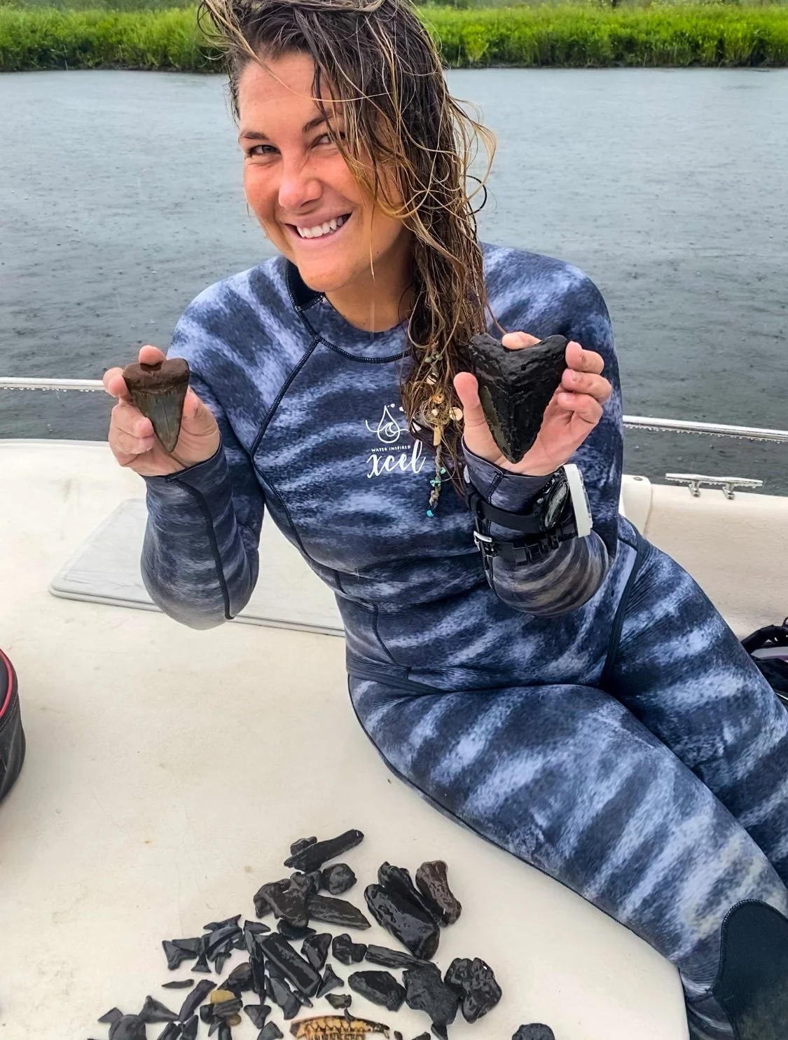 Woman in wetsuit holding two large black fossils on boat with fossils laid out on table in front of her, water and greenery in background.