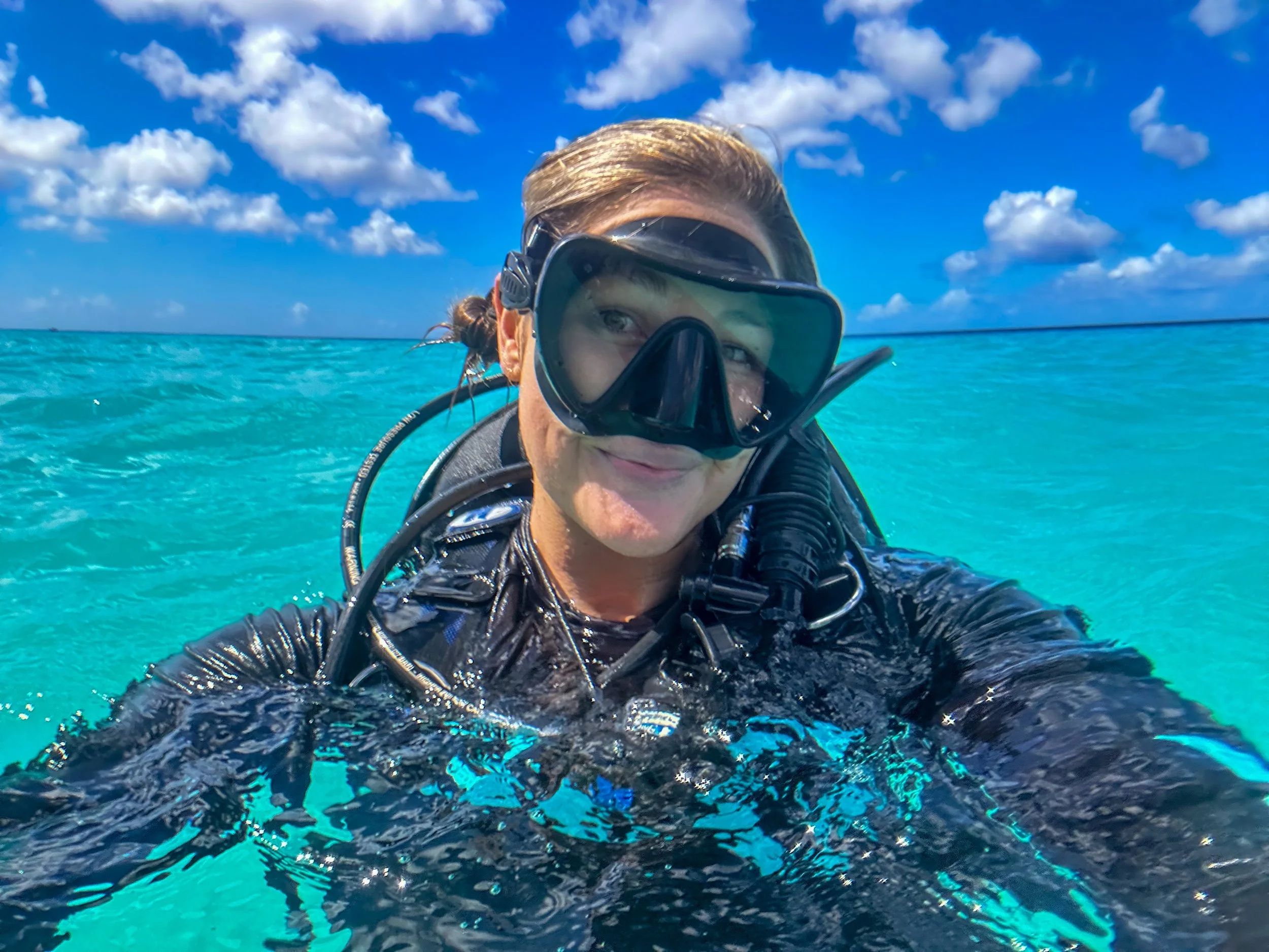 Person scuba diving in turquoise water, wearing a black wetsuit and diving mask under a partly cloudy sky.