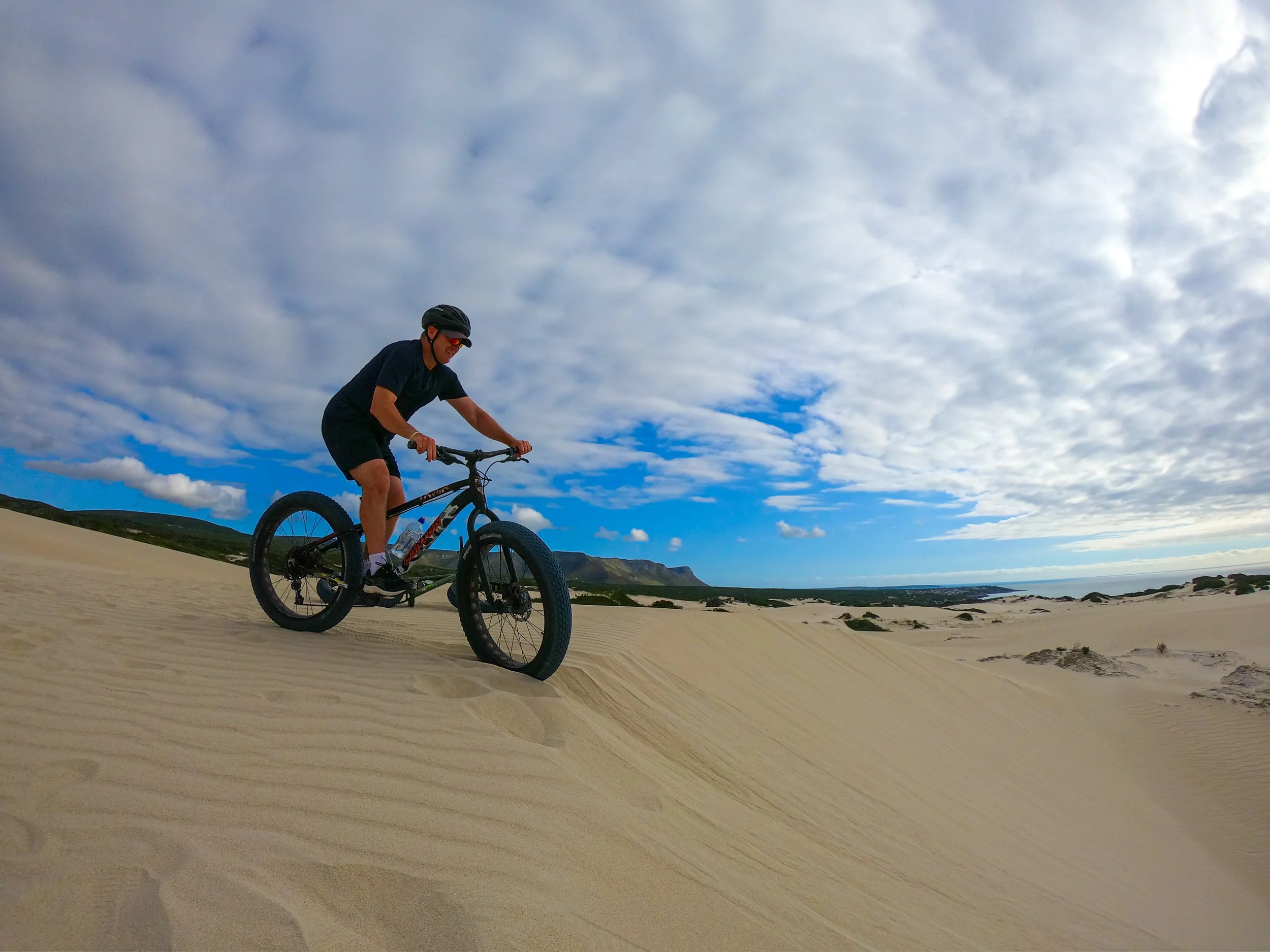 Person riding a mountain bike on sandy dunes under a partly cloudy sky.