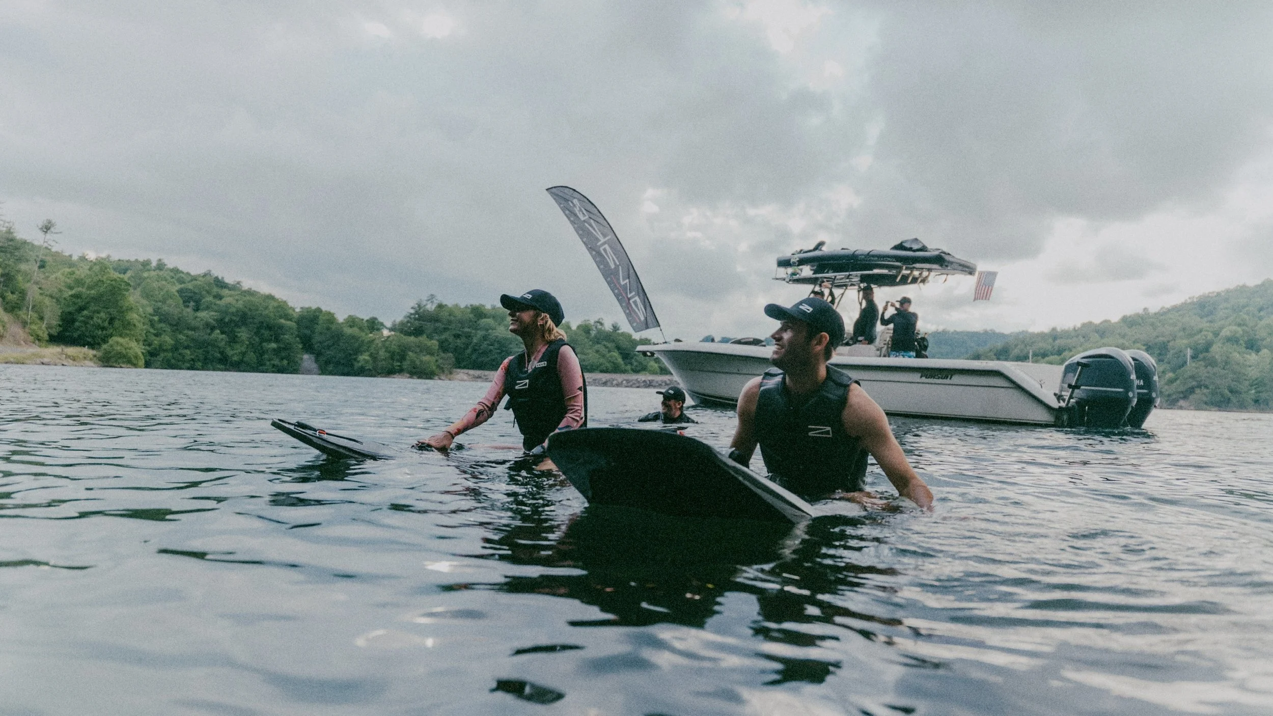 Two people in life jackets and caps stand in water next to wake surfboards, with a boat in the background on a cloudy day.