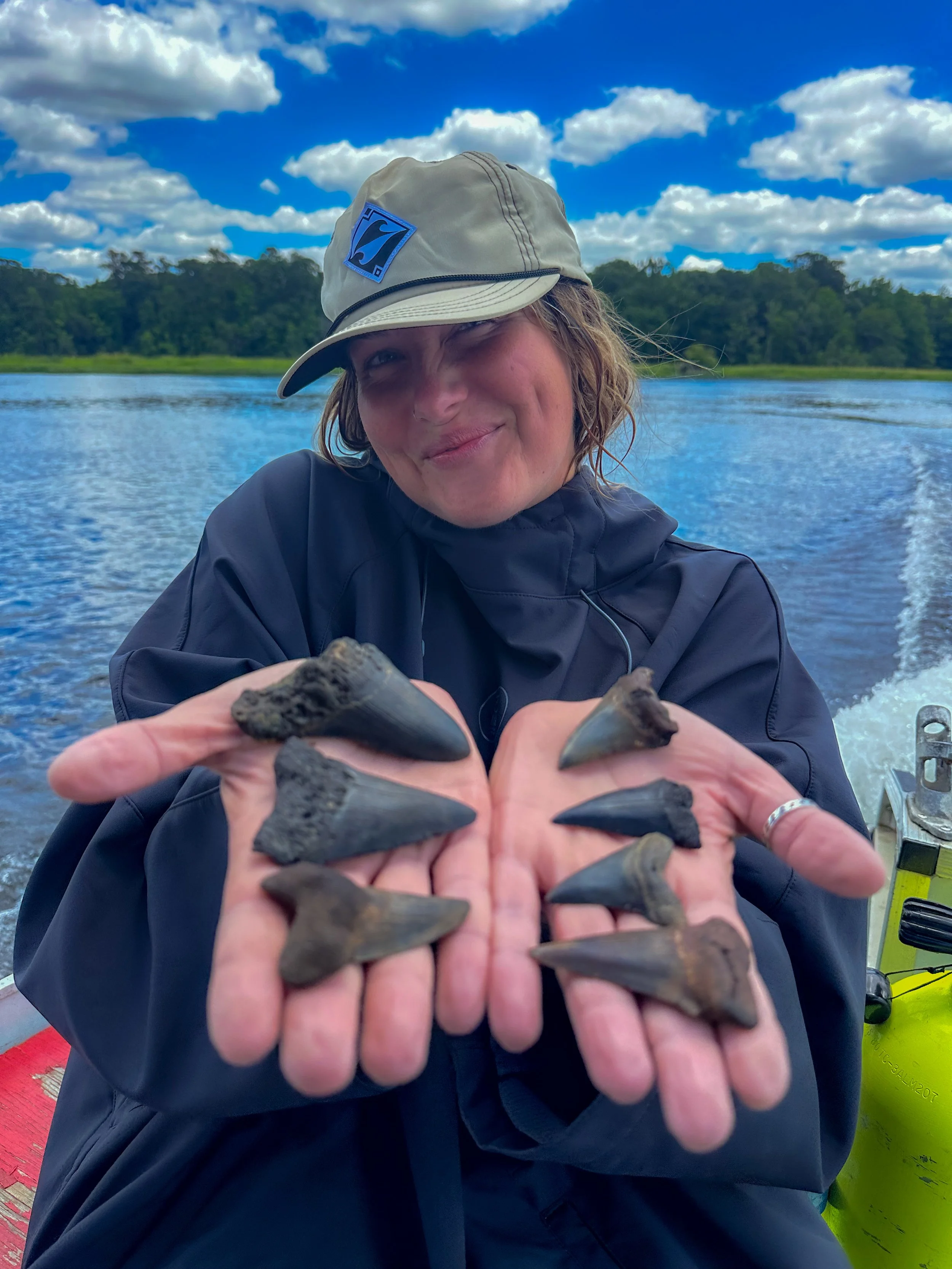 Person on a boat holding several fossilized shark teeth toward the camera with a lake and trees in the background.