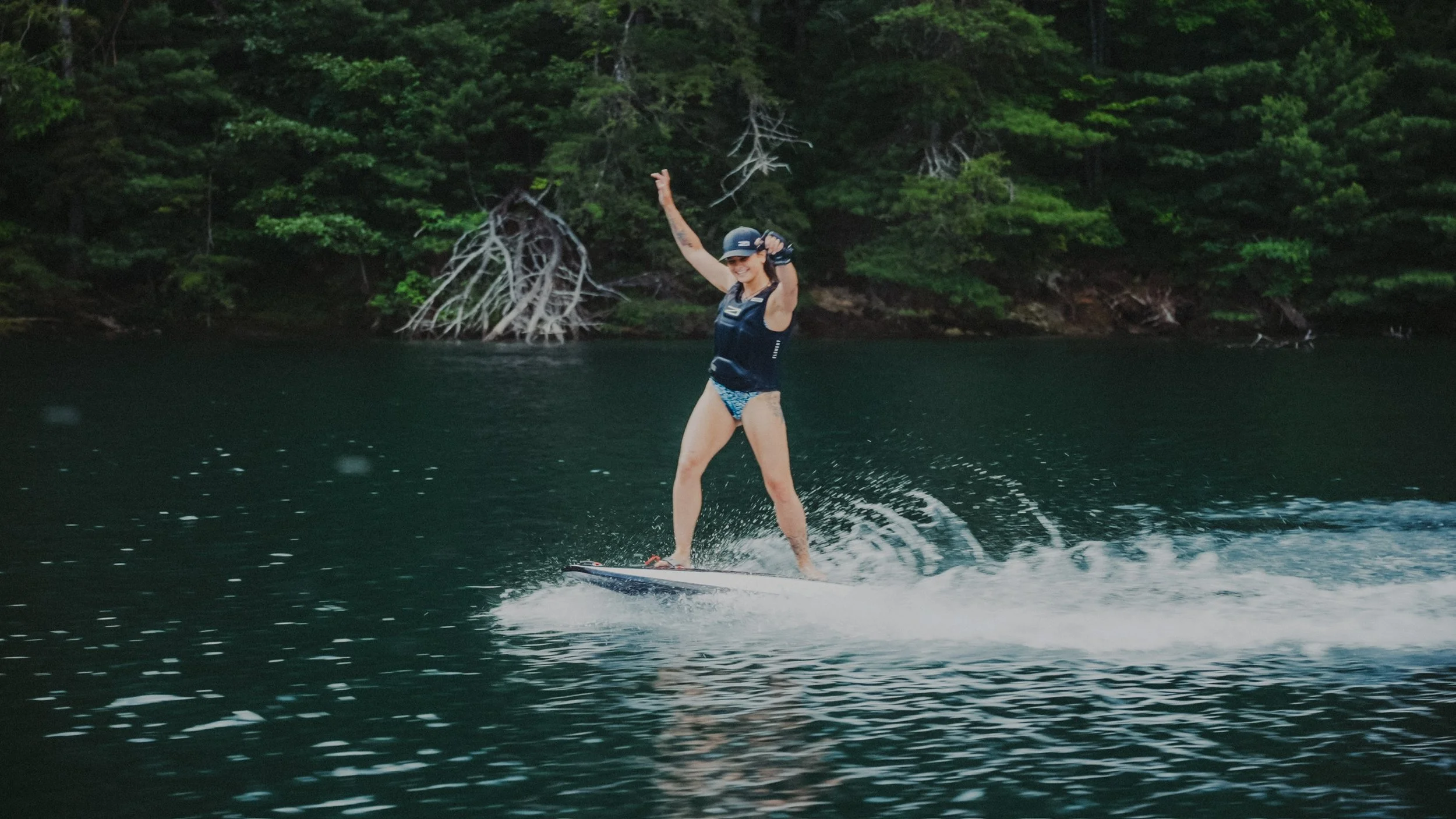 Woman wearing a helmet and life vest riding a wakeboard on a lake with green trees in the background.