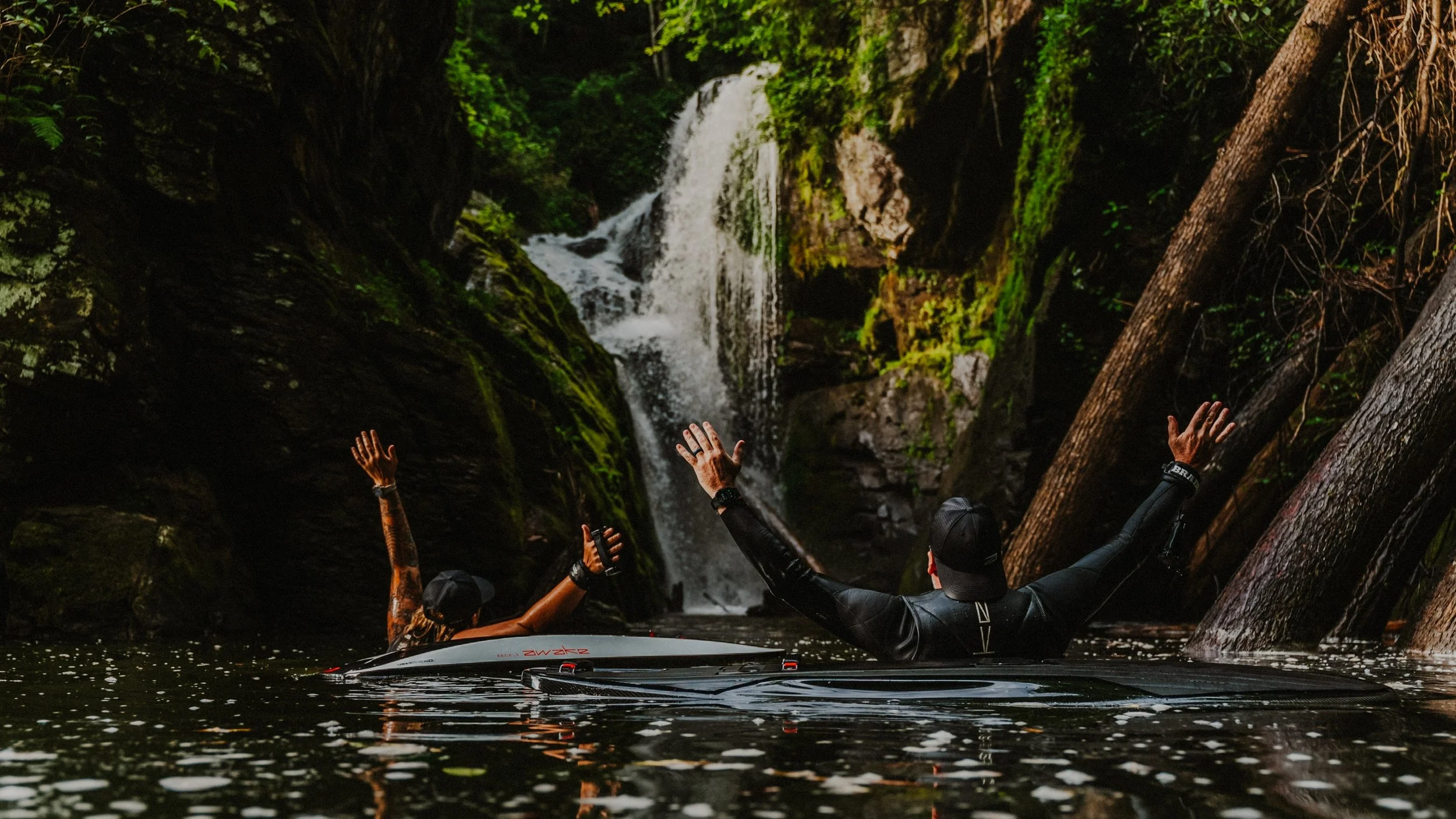 Two people on paddleboards raising their arms in front of a waterfall in a lush, green forest
