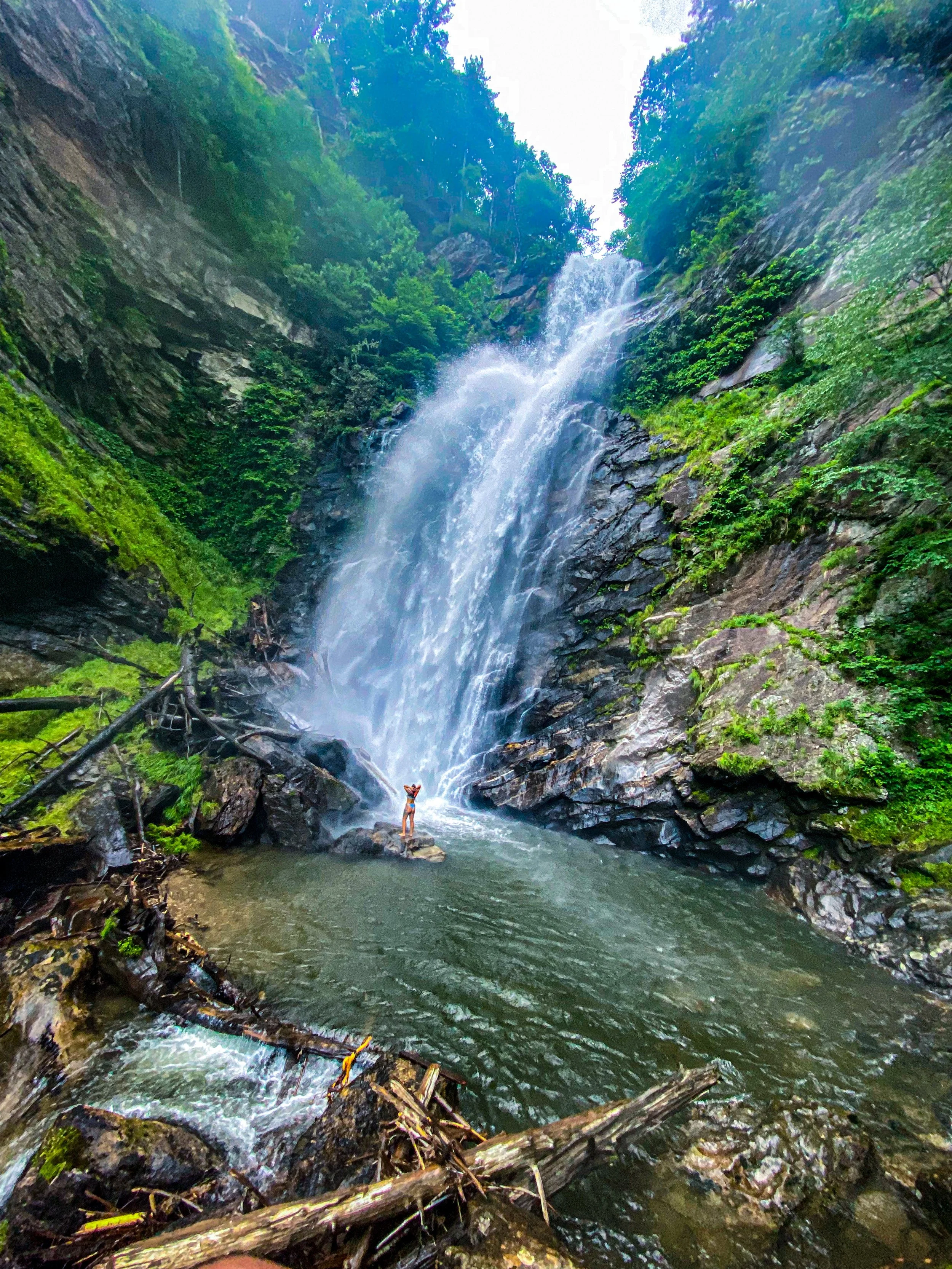 A person standing under a tall waterfall in a lush green forest, with rocks and water at the base.