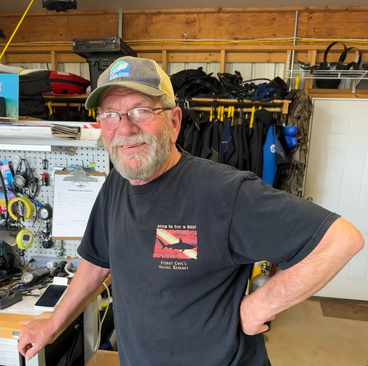 A smiling older man with glasses and a beard, wearing a baseball cap and a black t-shirt with a shark graphic, standing in a workshop or gear storage area with diving equipment and tools in the background.